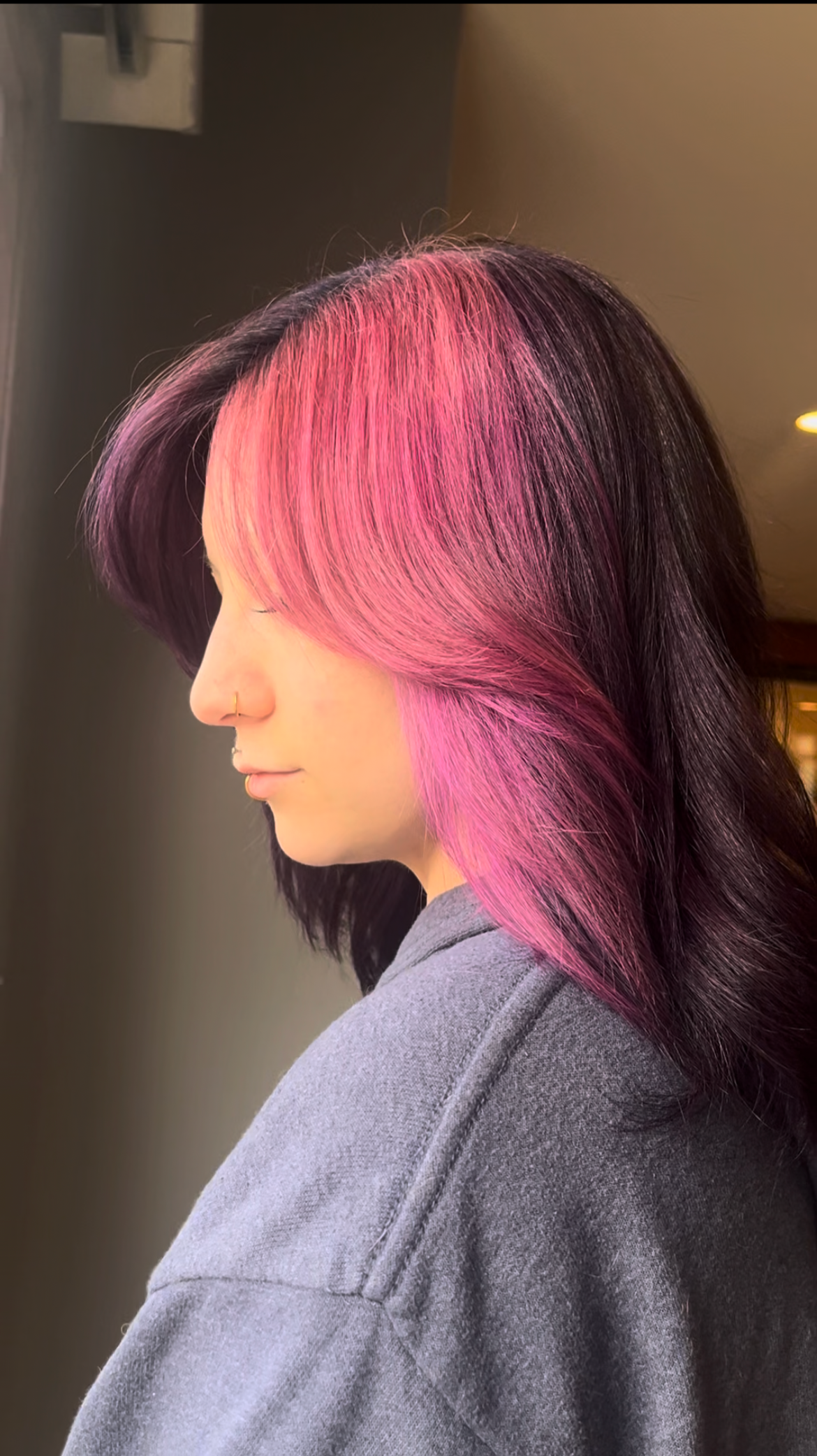 Side profile of a woman with pink and purple layered hair, wearing a gray sweatshirt, and a small nose ring.