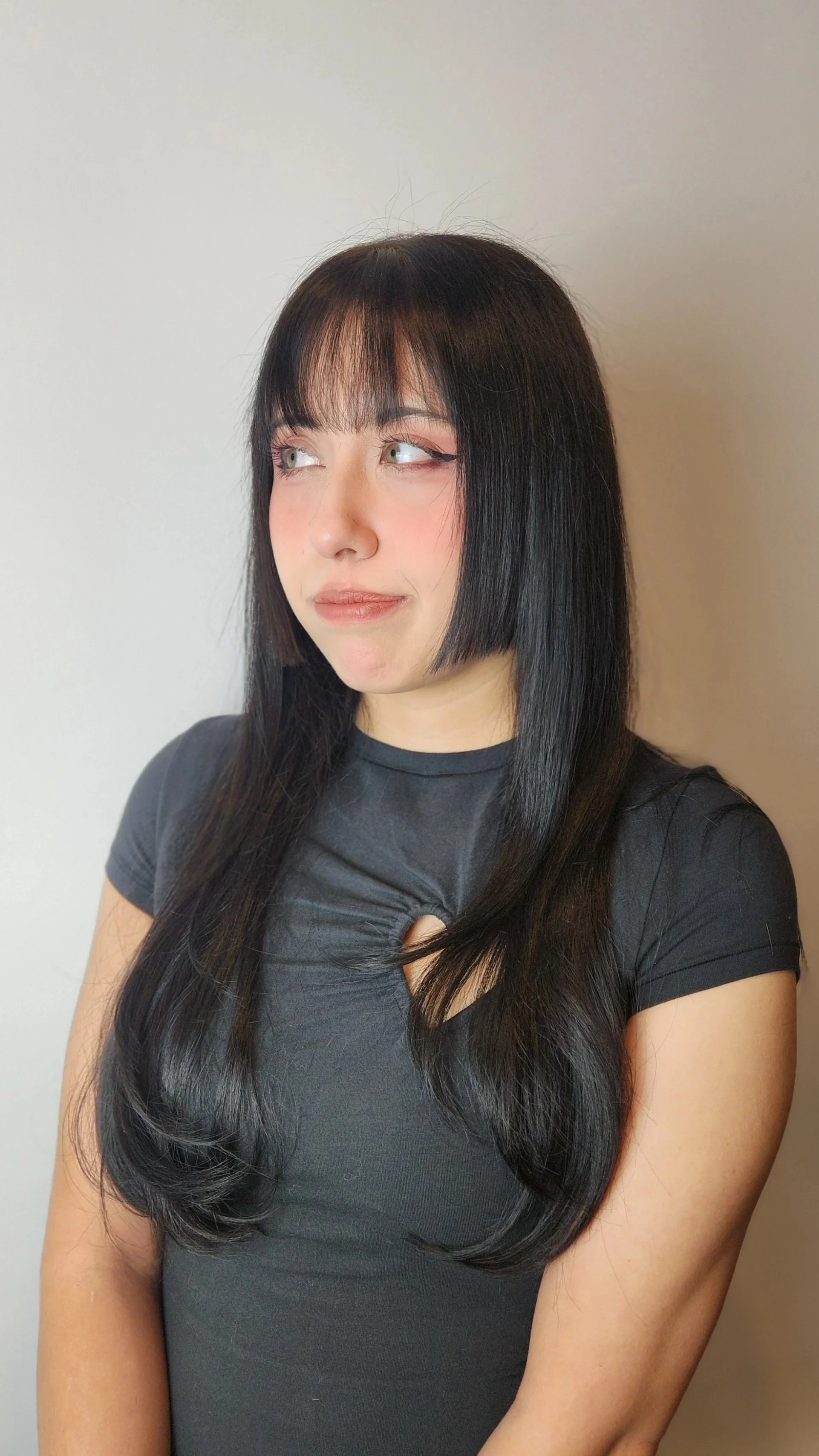 A young woman with long black hair and bangs, wearing a black t-shirt with a cut-out detail, looking to her left against a plain white wall background.