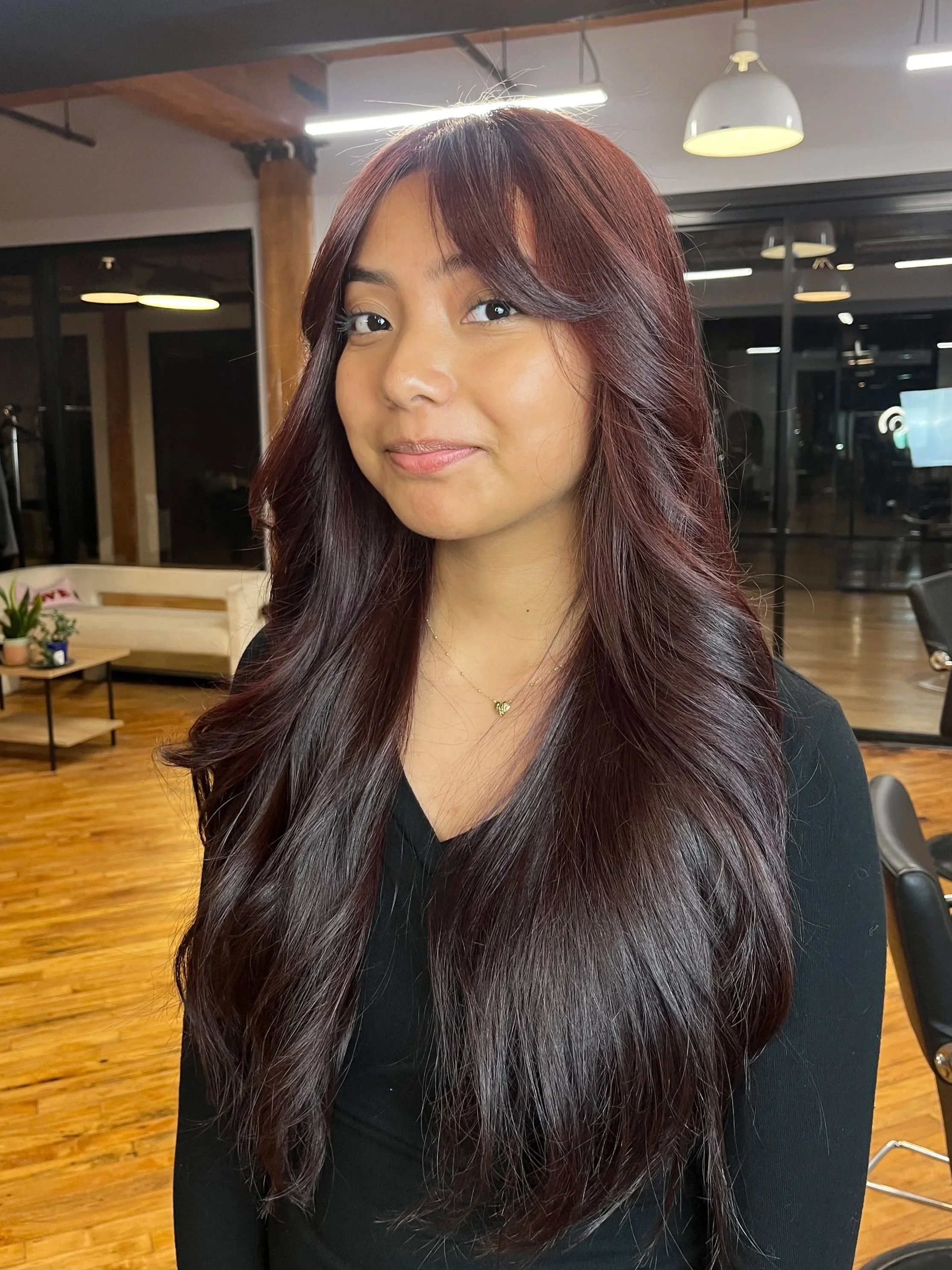 A young woman with long, wavy, dark red hair, wearing a black top and a delicate gold necklace, standing in a modern indoor space with wooden flooring, large windows, and contemporary furniture.