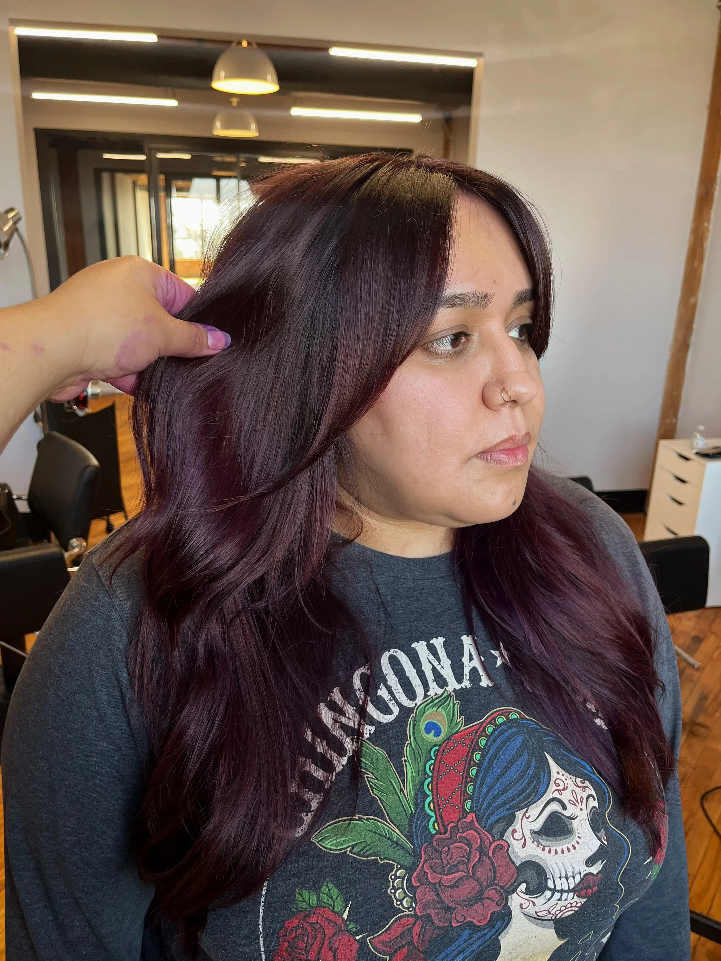 A woman with long, wavy purple hair sitting in a hair salon, having her hair styled by a hairstylist.