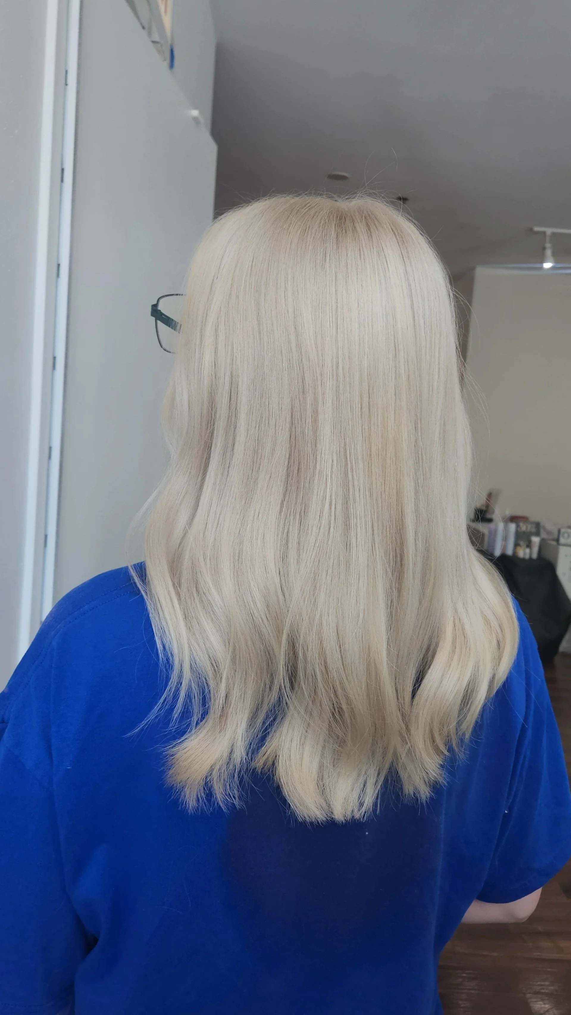 Back of a woman's head with long, wavy platinum blonde hair, wearing a blue shirt.