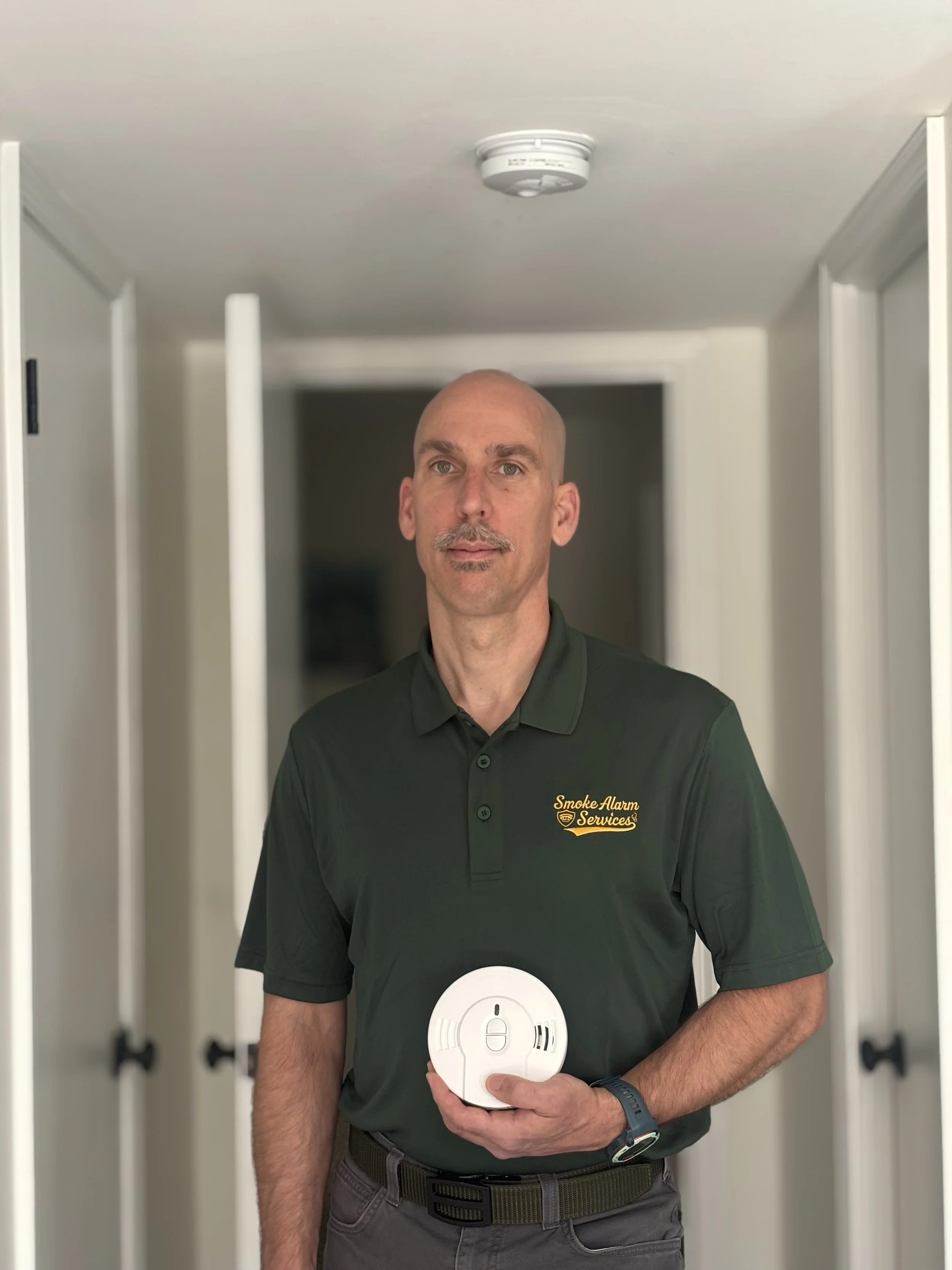 Man wearing a dark green polo shirt with 'Smoke Alarm Services' logo, holding a smoke detector in front of an open smoke alarm system in a hallway.