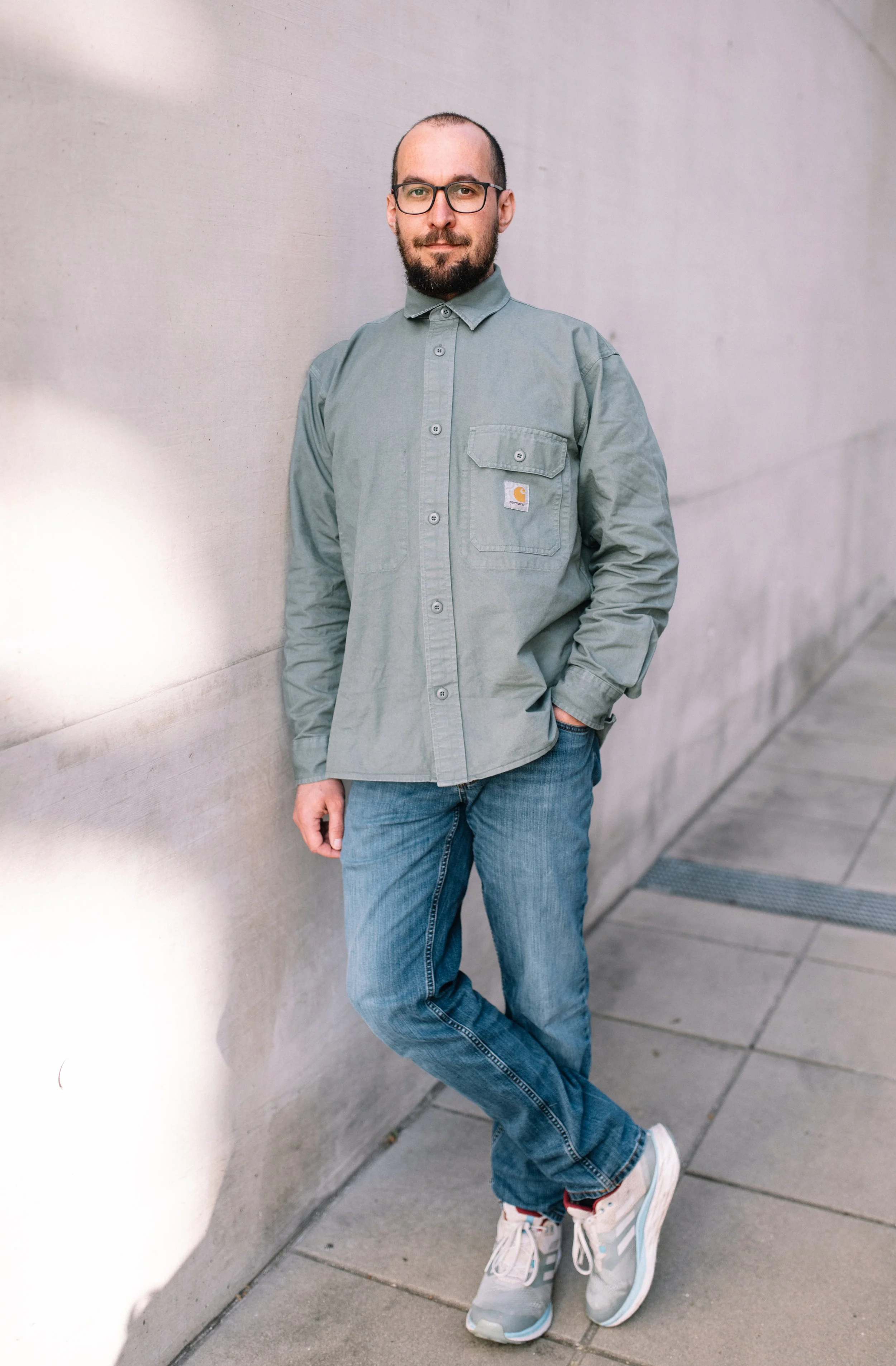 Ein junger Mann mit Brille, Bart und kurzem Haar steht entspannt an einer Betonwand in einer urbanen Umgebung, trägt eine graue Carhartt-Jacke, blaue Jeans und weiße Sneaker.