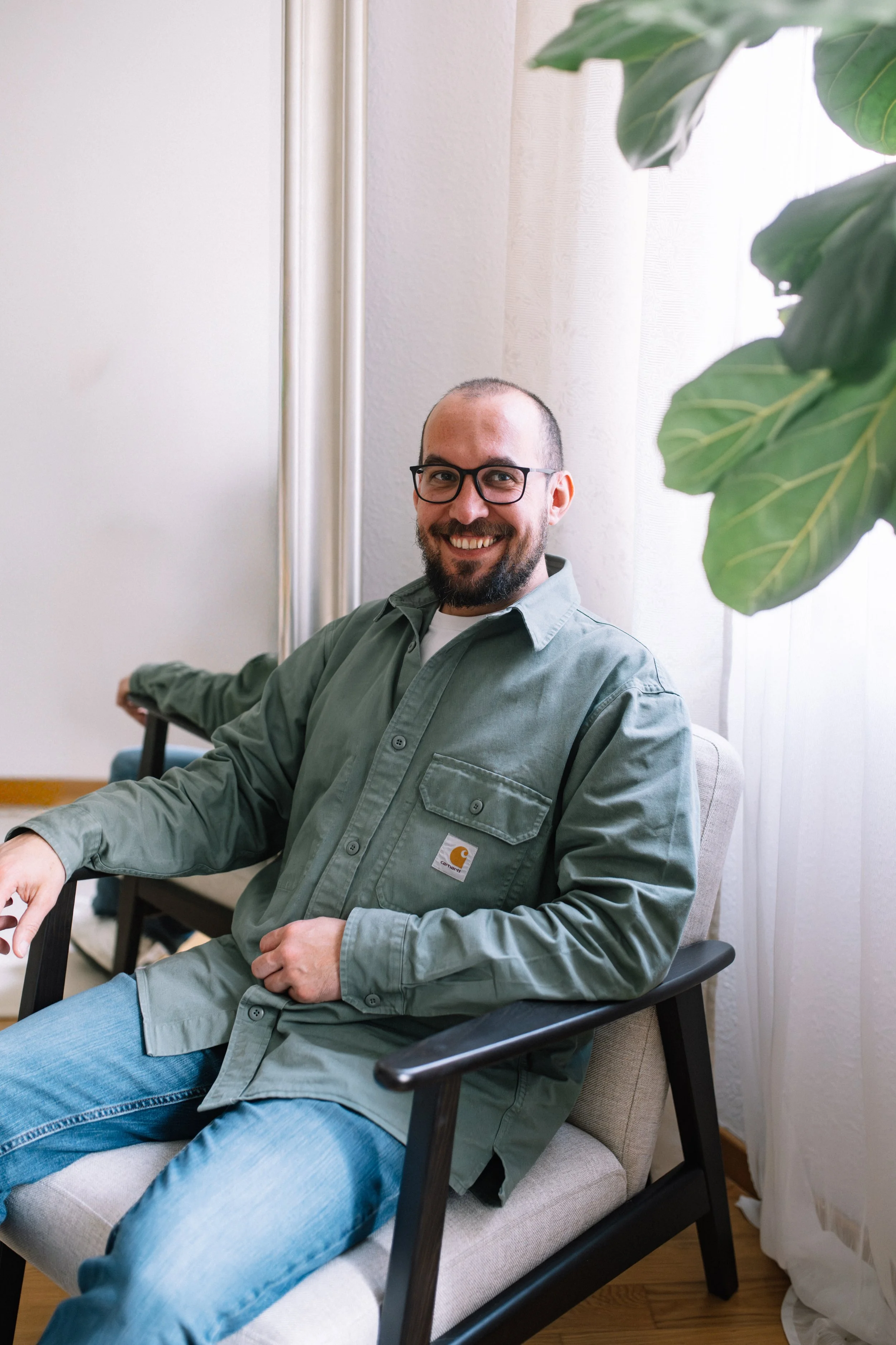 Ein lächelnder Mann mit Brille sitzt entspannt auf einem Stuhl in einem Wohnzimmer neben einer Zimmerpflanze.