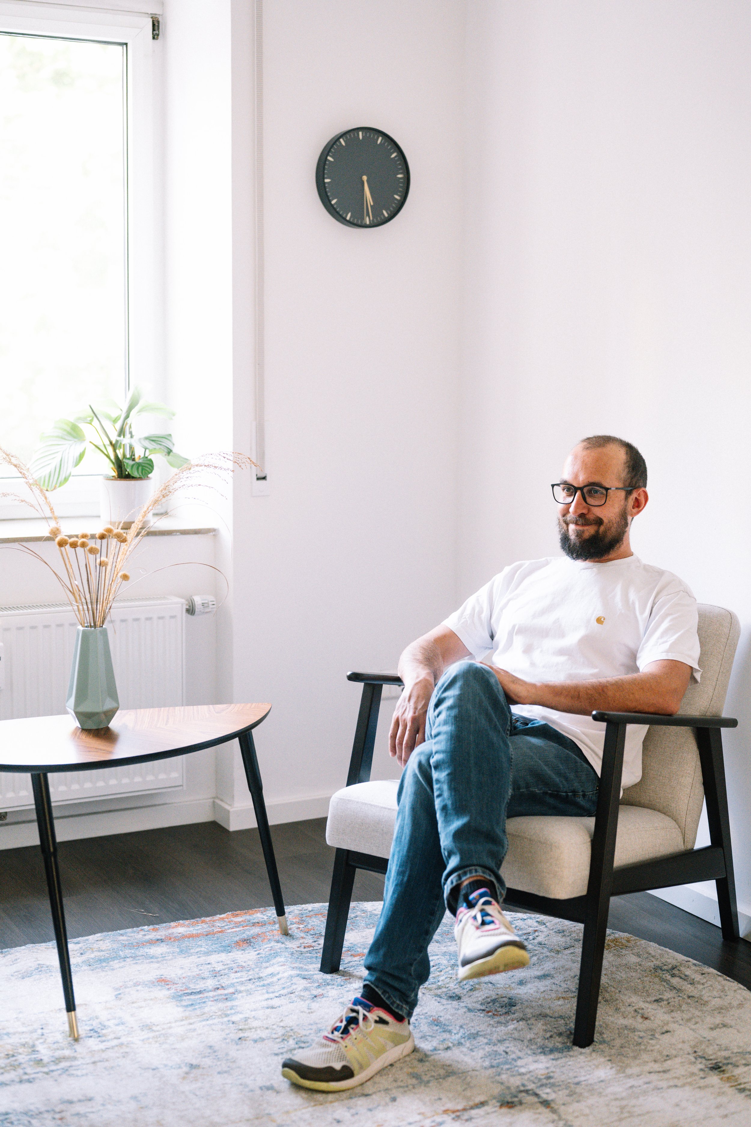 Ein Mann sitzt entspannt in einem Wohnzimmer auf einem beige-grauen Sessel, trägt eine weiße T-Shirt, Jeans und Turnschuhe, vor einem Fenster mit Pflanzen und einer schwarzen Wanduhr.