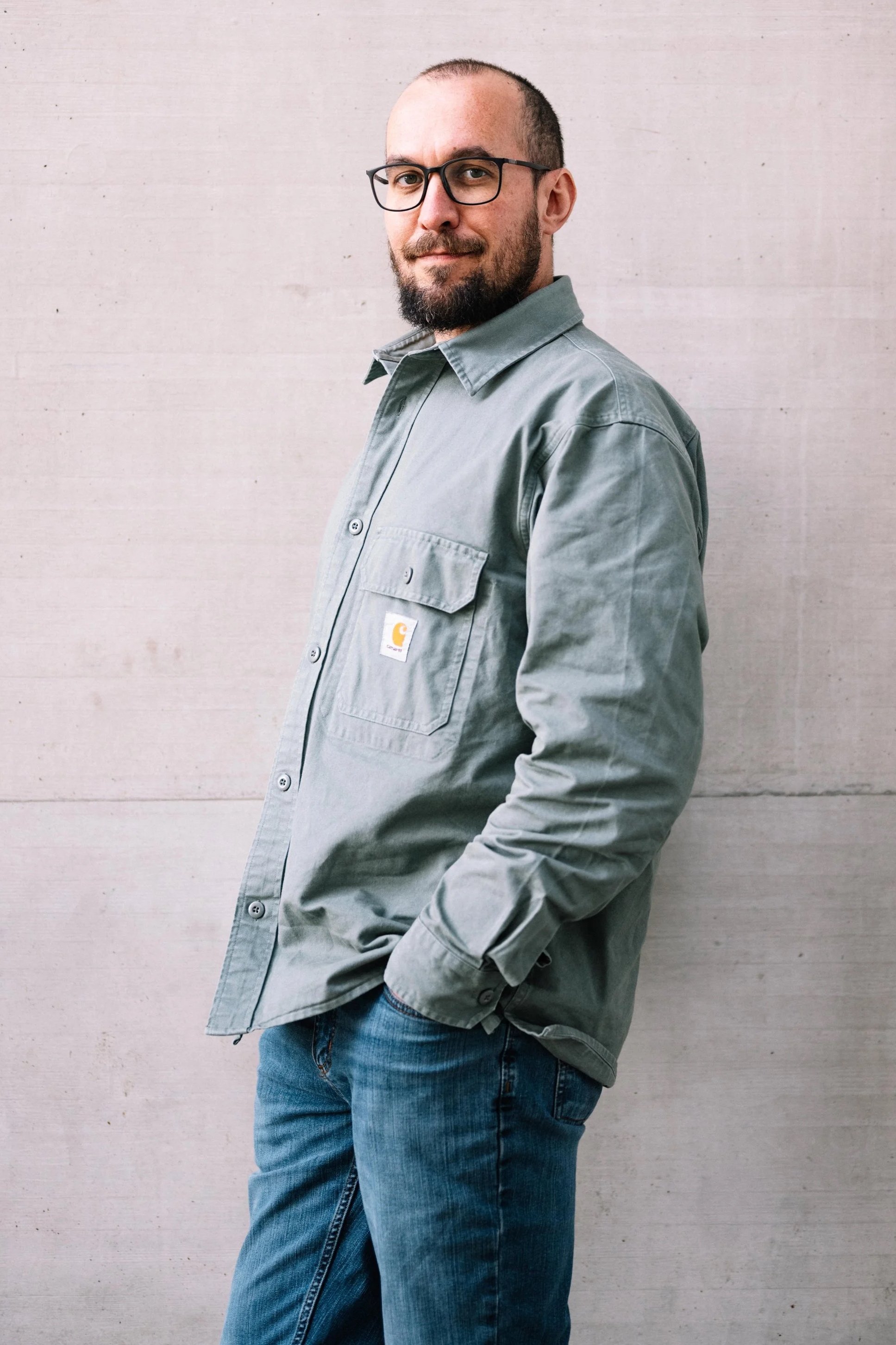 Ein Mann trägt eine graue Jacke und eine Brille, posiert vor einer hellen Holzwand.