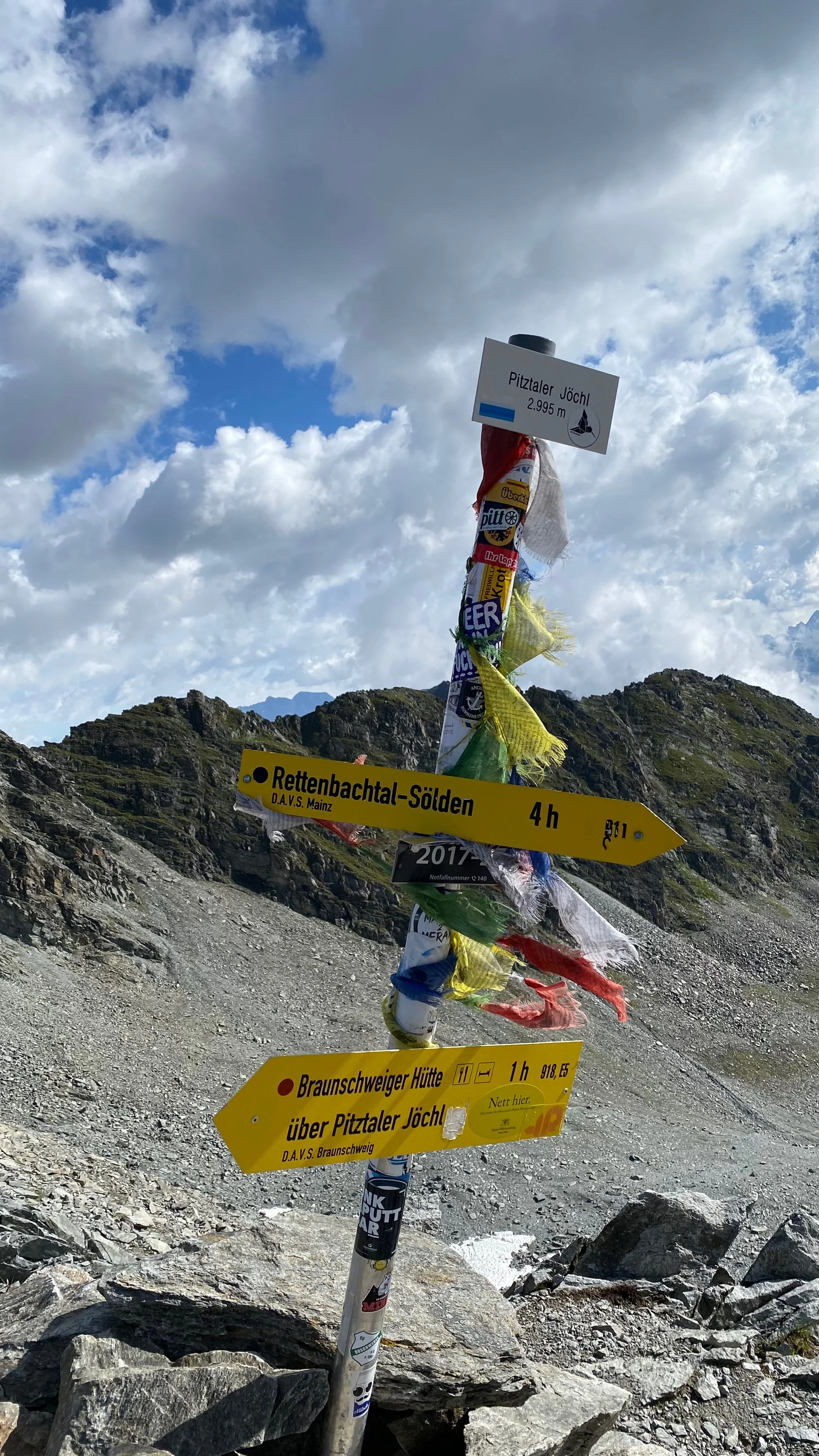 Wegweiser auf einem Bergpfad mit Richtungshinweisen zu Rettentbachtal-Sölden und Braunschweiger Hütte, in den Bergen bei bewölktem Himmel.