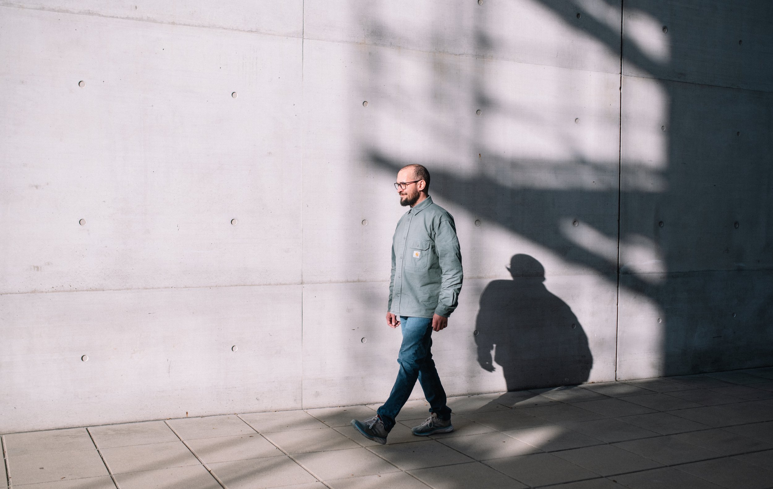 Ein Mann mit Brille, grauer Jacke und blauen Jeans geht an einer weißen Betonwand entlang. Sein Schatten und der Schatten eines Baumes sind auf der Wand sichtbar.