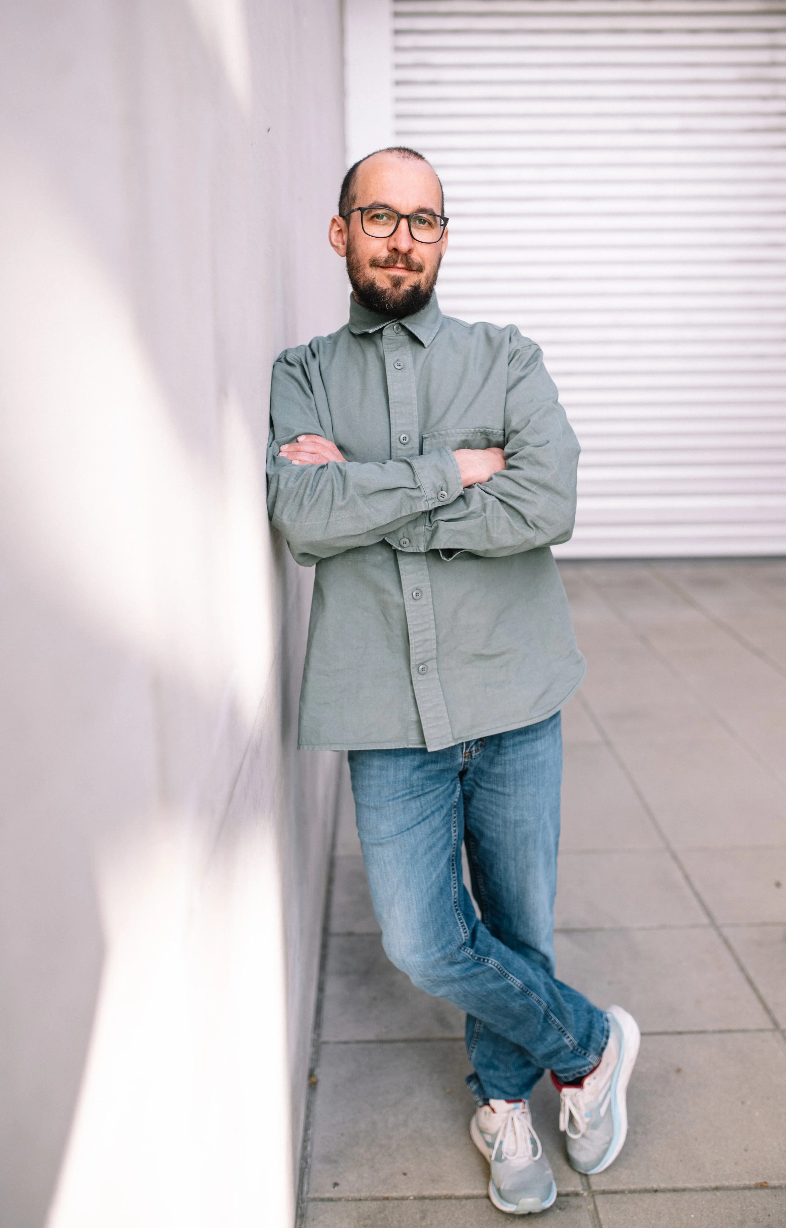 Ein Mann mit Brille, Bart und kurzhaarigem Kopf lehnt an einer weißen Mauer, trägt eine graue Hemd und blaue Jeans, und steht auf einem Gehweg.