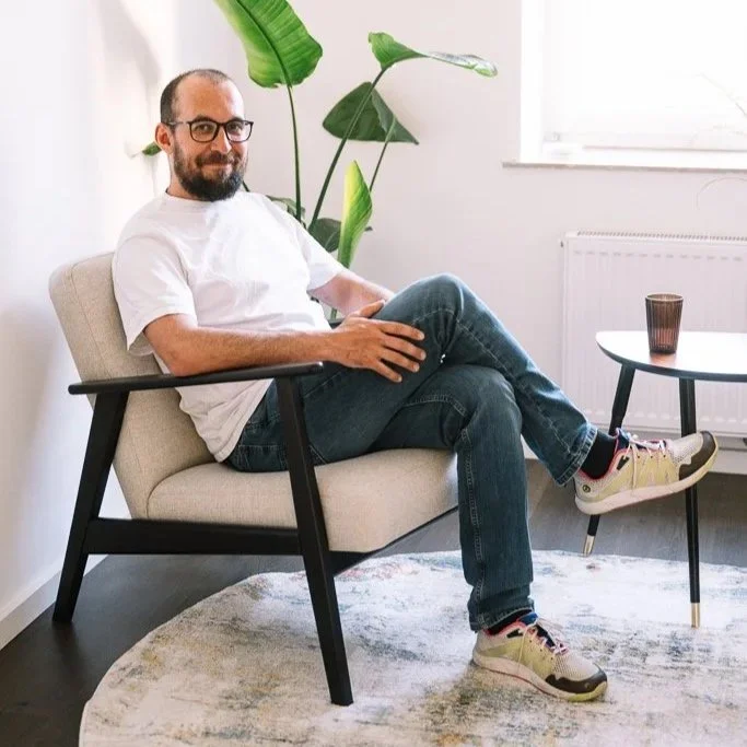 Ein Mann sitzt entspannt in einem cremefarbenen Sessel, trägt Brille, weißes T-Shirt, Jeans und Turnschuhe, im Hintergrund große grüne Pflanzen und ein Fenster, neben einem kleinen Tisch mit einem Glas. Modern eingerichteter Raum.