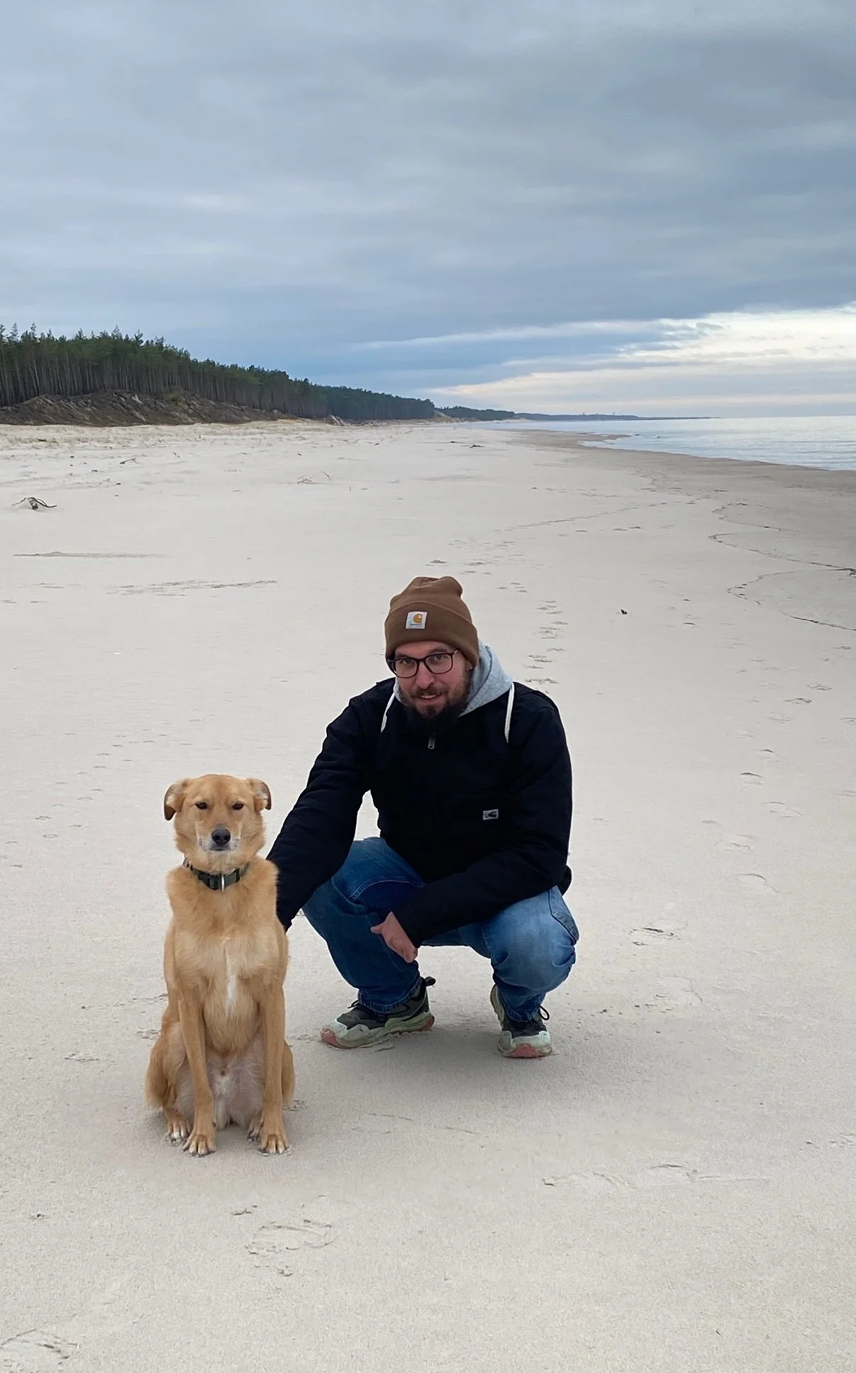 Ein Mann mit Brille, braunem Mützen, schwarzer Jacke, blauer Jeans und Sneakers kniet an einem Hund auf einem verlassenen Sandstrand bei bewölktem Himmel.