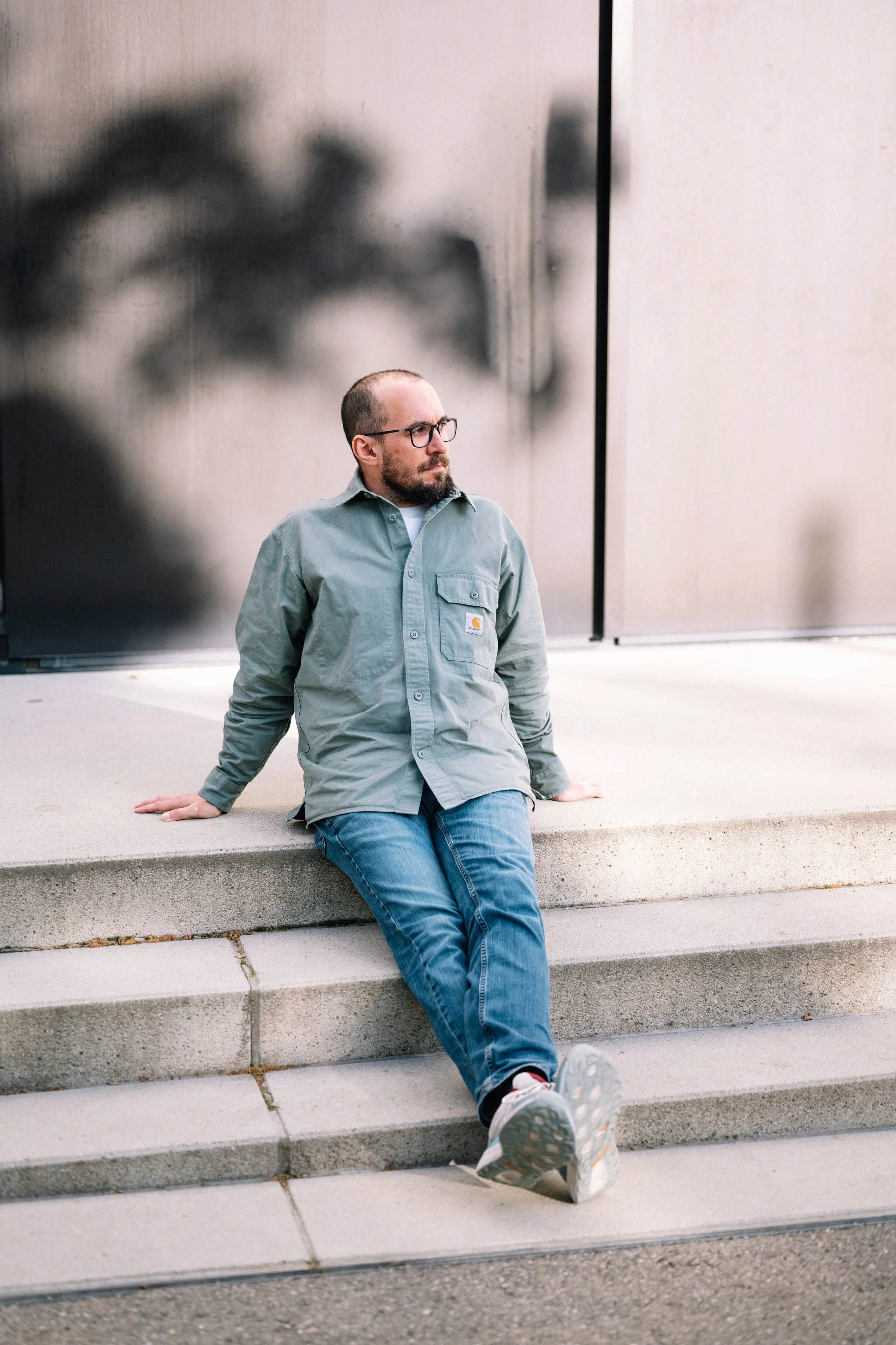 Mann mit Brille, kurzhaarig, in grauem Hemd und blauen Jeans, sitzt auf Stufen vor einem modernen Gebäude mit Metallfassade, Blick nach links gerichtet.
