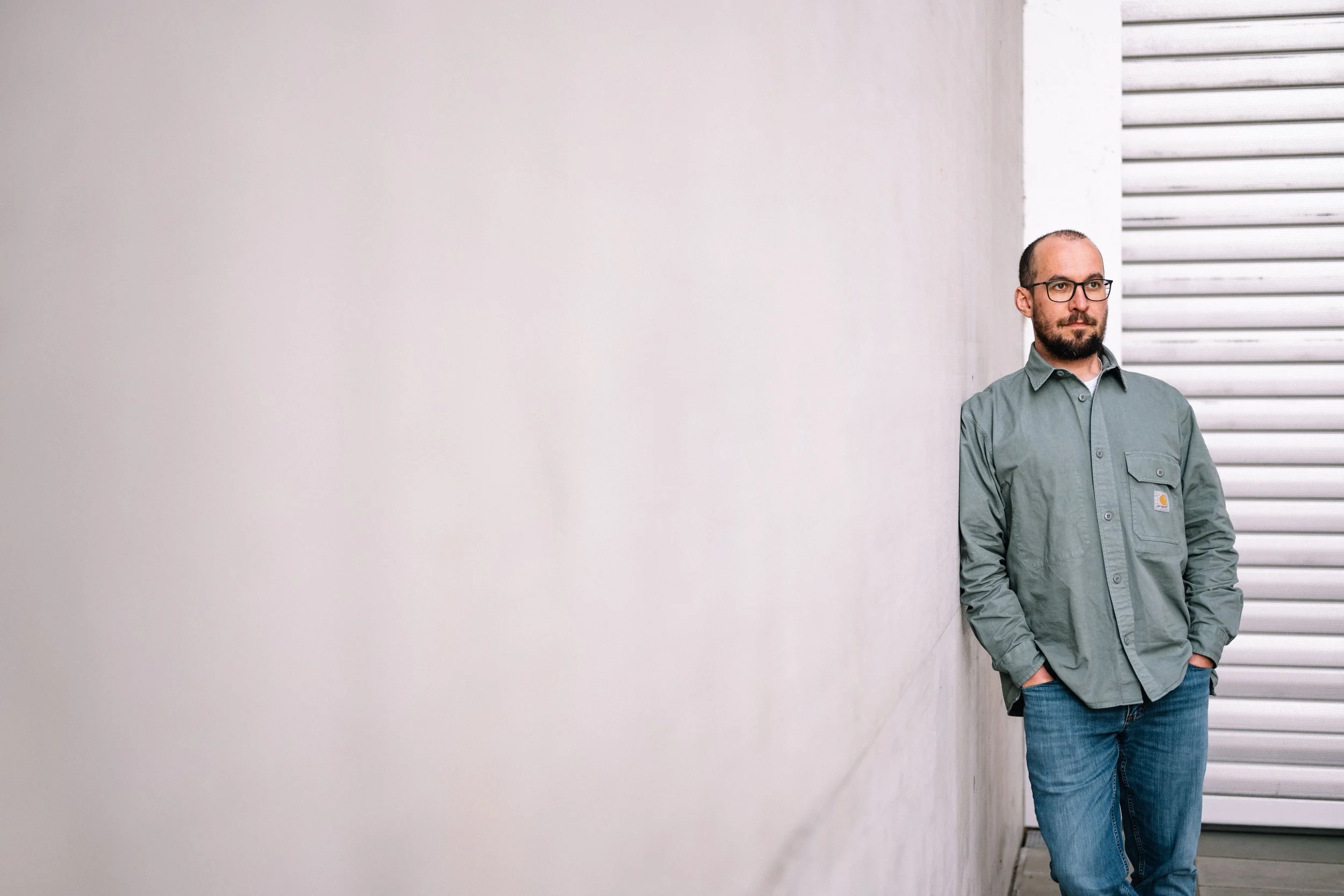 Ein Mann mit Brille trägt eine grüne Jacke und blaue Jeans, steht an einer weißen Wand und blickt nach rechts.