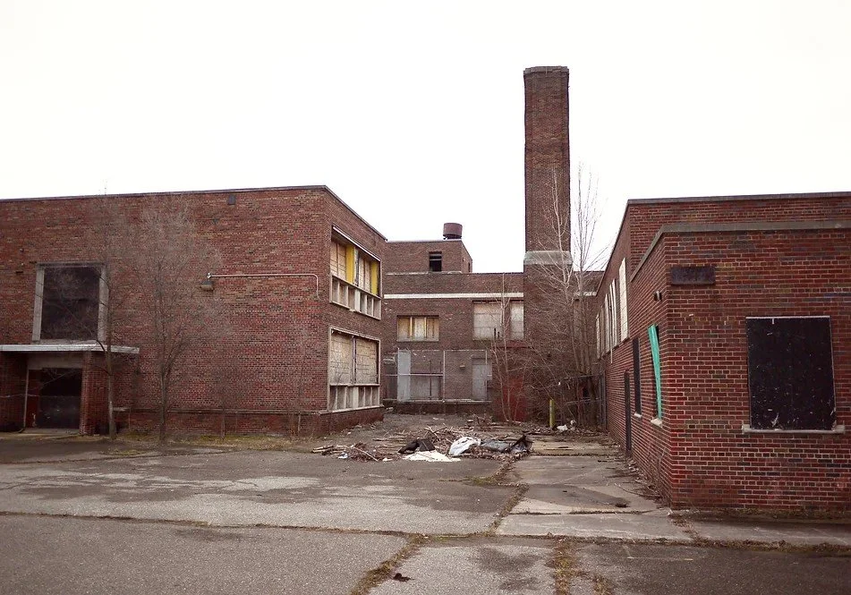 Abandoned and dilapidated brick building with boarded-up windows and overgrown trees, debris on cracked pavement in a run-down area.
