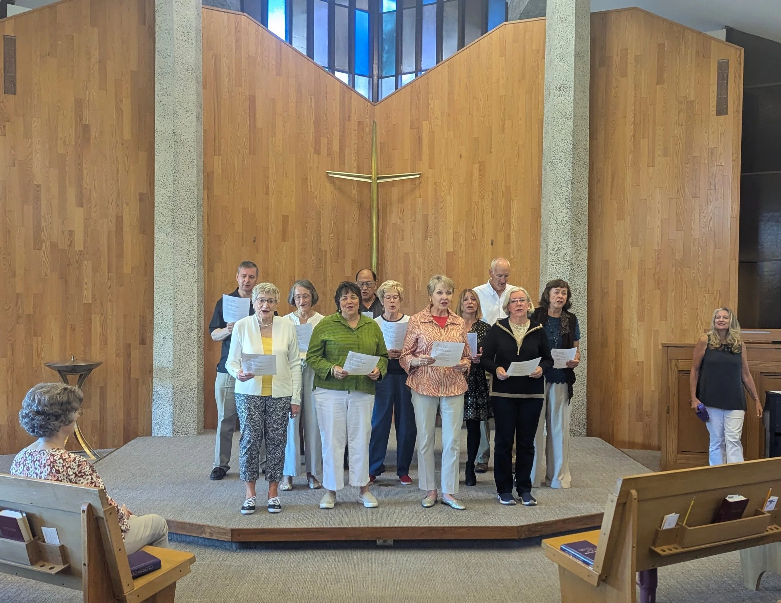 A group of people singing in a church during a worship service, standing on a small elevated platform with song sheets in hand, behind a large wooden cross on the wall.