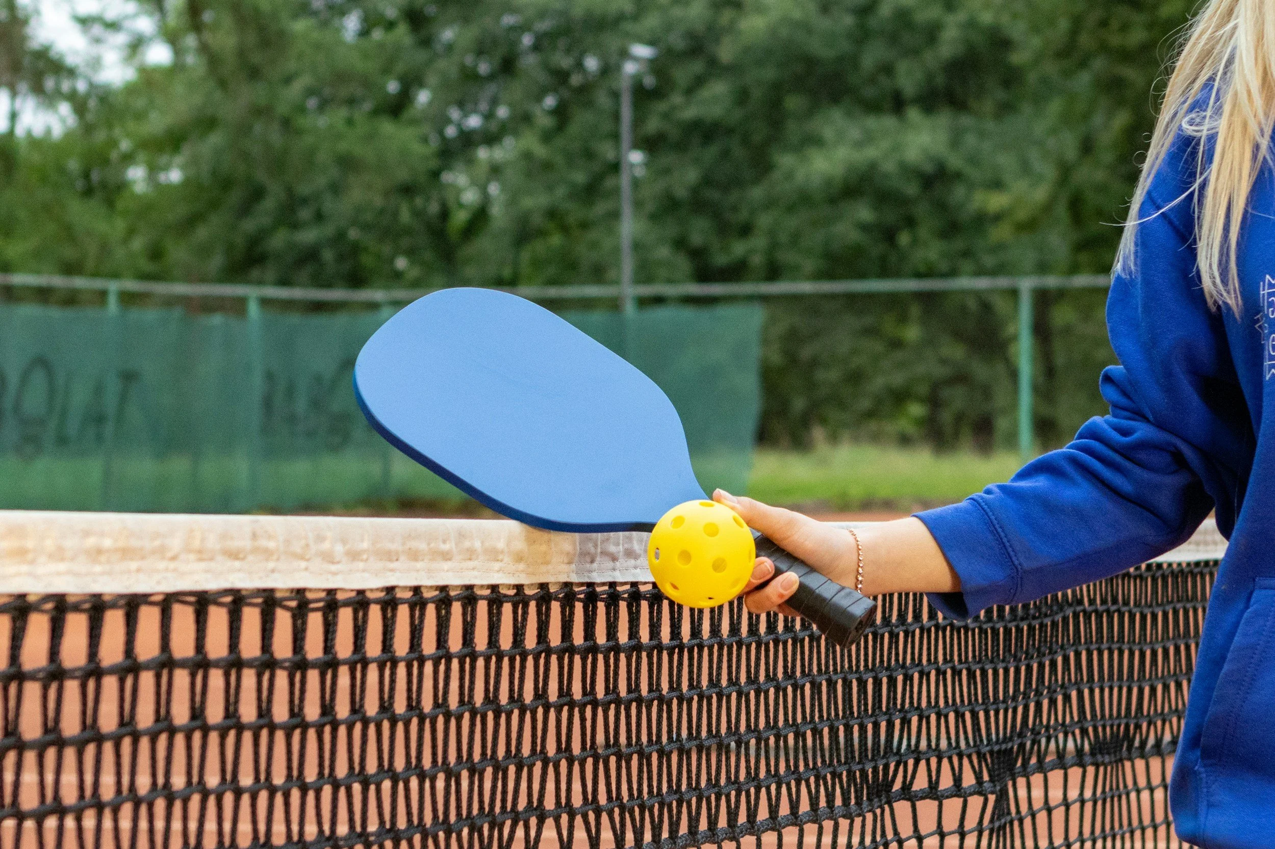 Close-up of a woman playing pickleball, holding a blue paddle and a yellow perforated ball, standing behind a black net on an outdoor tennis court with green trees in the background.