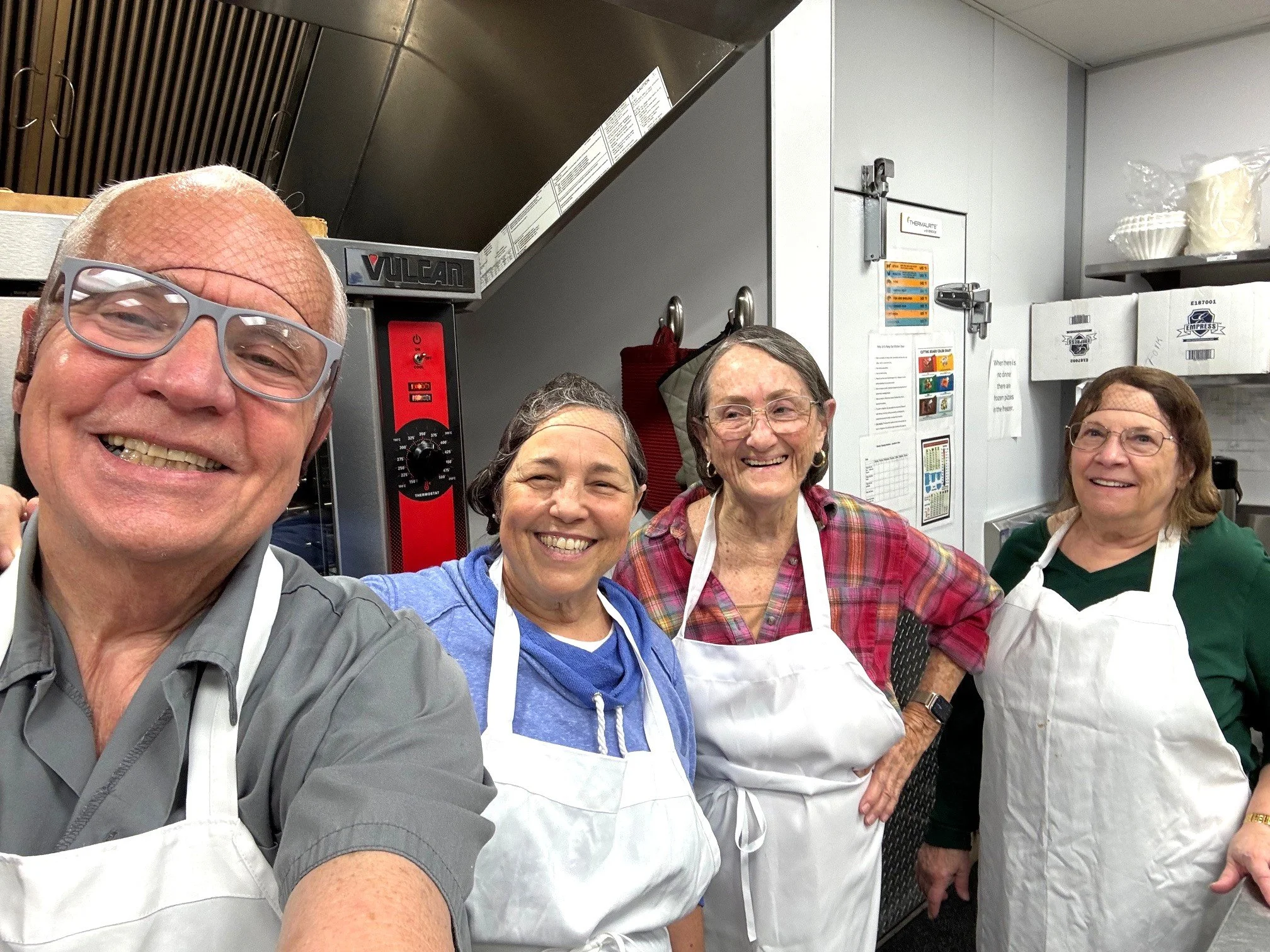 Group of four smiling elderly people wearing aprons in a kitchen setting.