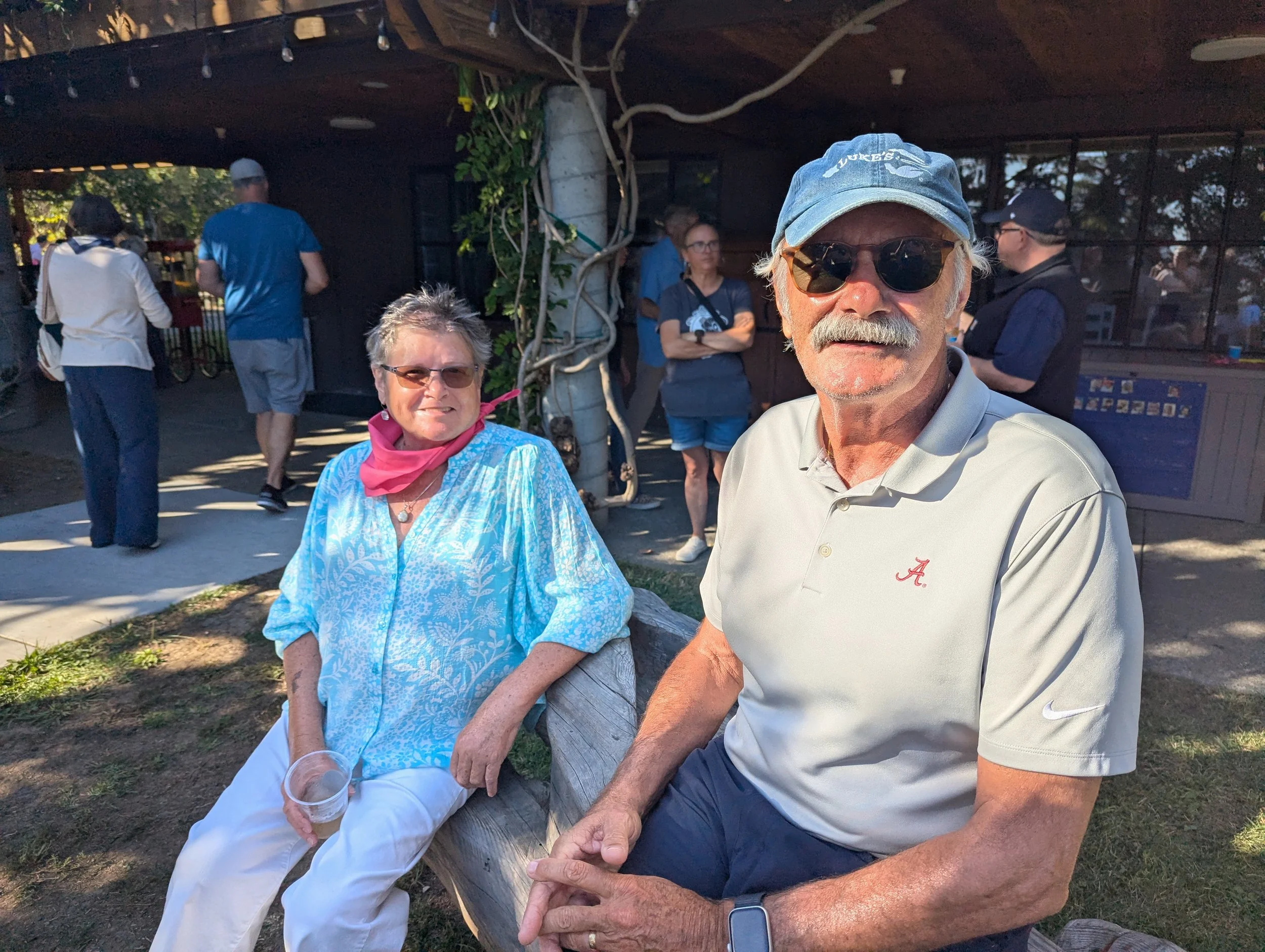 A man wearing sunglasses, a blue cap, and a gray polo shirt is sitting next to a woman in a blue patterned top with a pink scarf, both smiling. They are outdoors in a park with other people in the background.