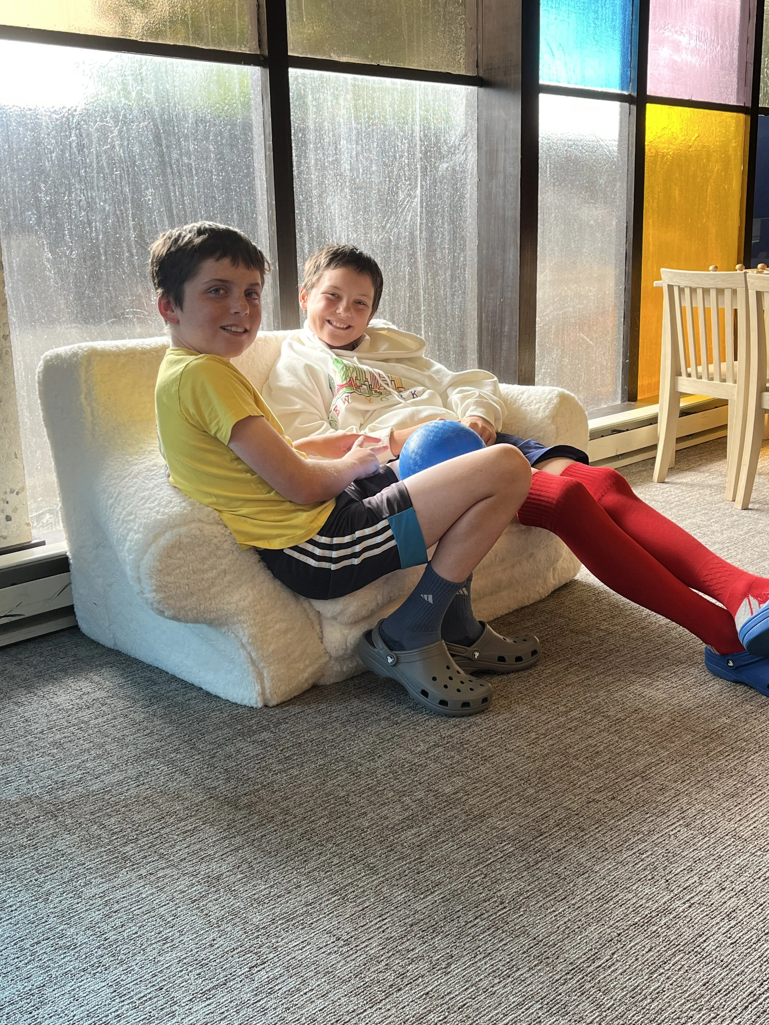 Two boys sitting on a white plush armchair in front of a large window with colorful stained glass panels. One boy is wearing a yellow t-shirt and black shorts, the other is in a white hoodie and black shorts. They are smiling and holding a blue ball.