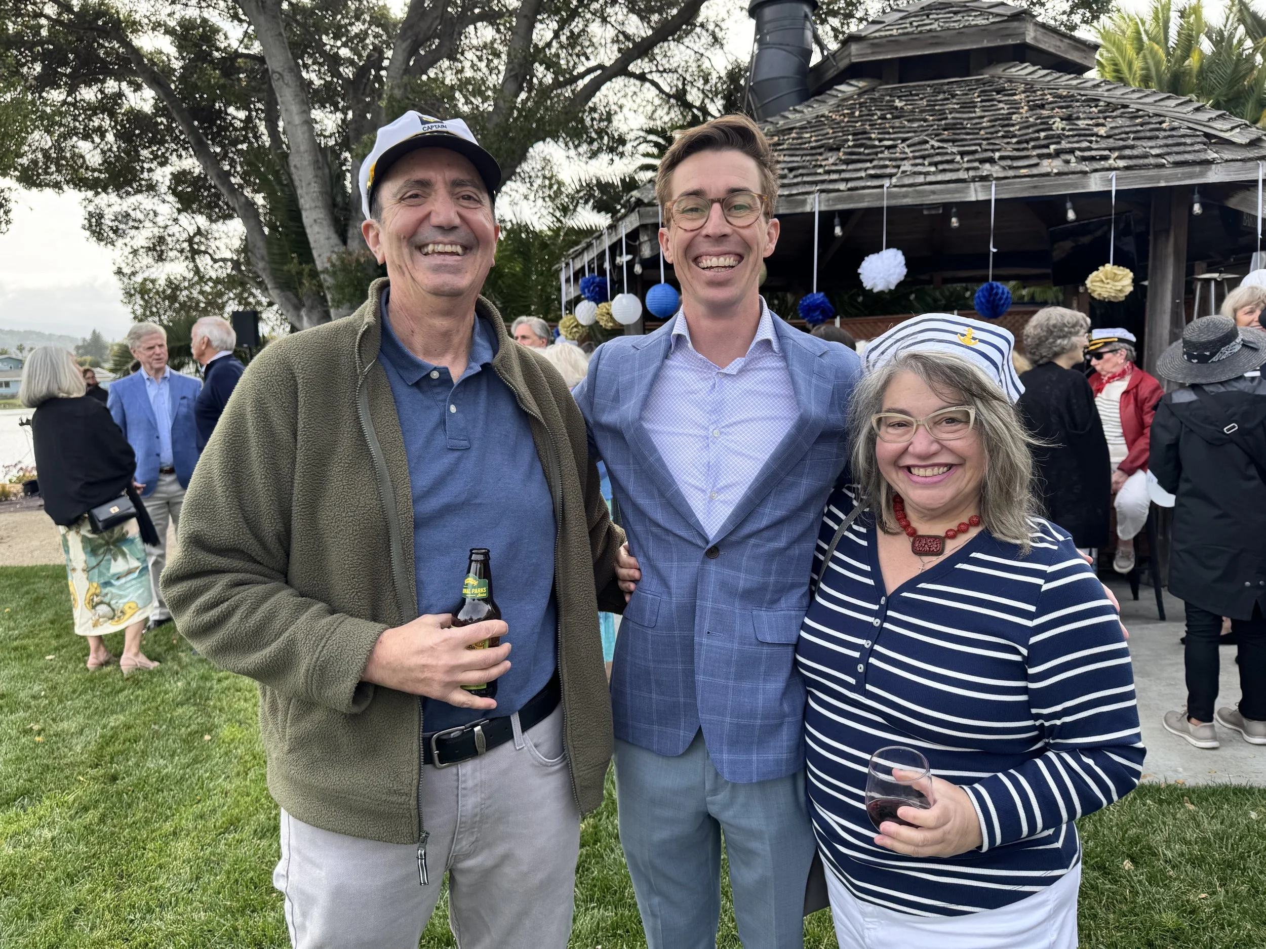Three people smiling at an outdoor gathering, standing close together. Behind them, other guests are mingling near a wooden pavilion decorated with blue, white, and yellow pom-poms and lanterns. The setting appears festive with a mix of casual and se