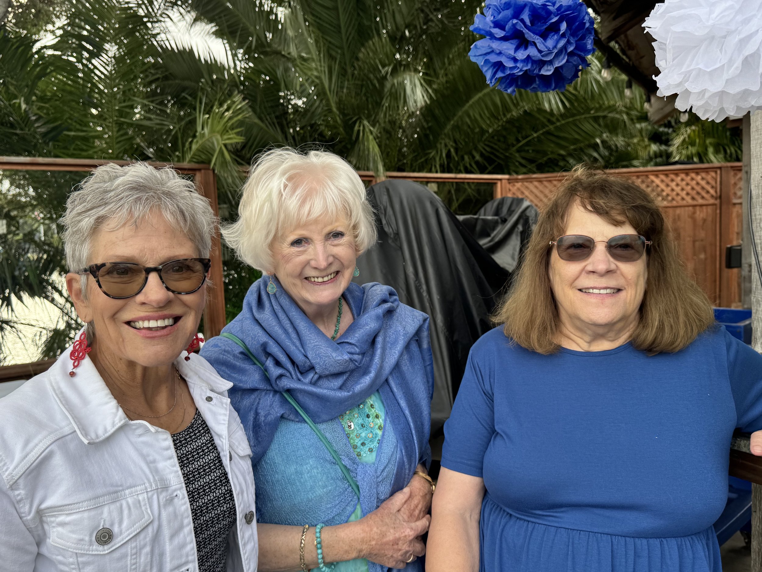 Three smiling women gathering outdoors, with tropical palm trees and blue and white paper decorations in the background.