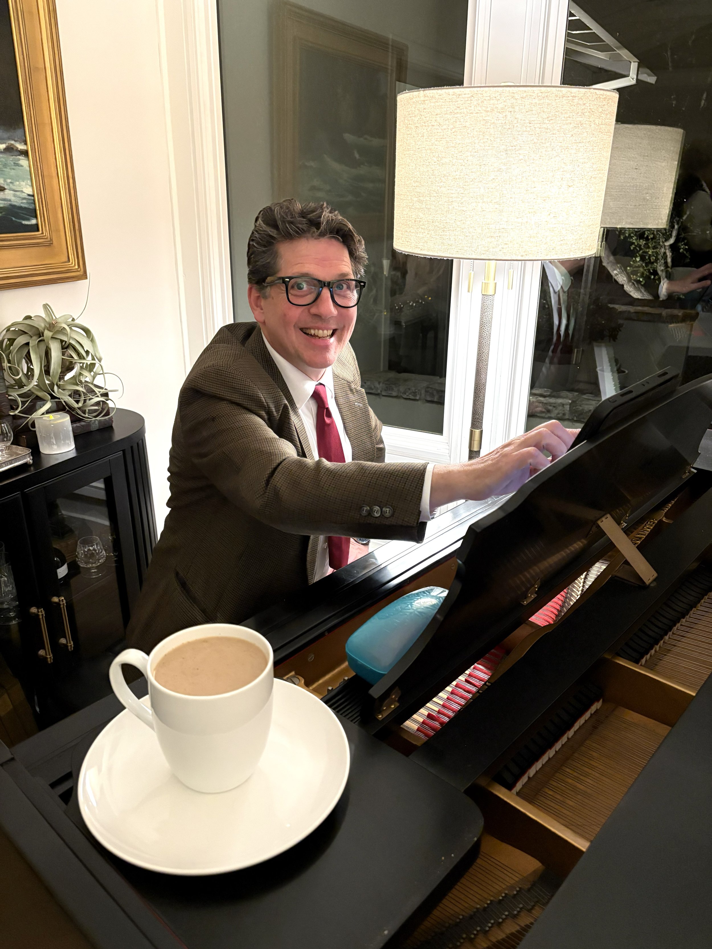Man wearing glasses, smiling, sitting at a piano, with a cup of coffee on a white saucer in the foreground.