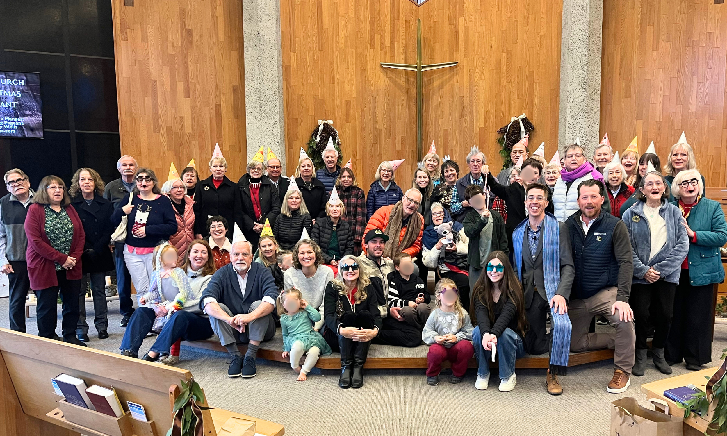 A large group of people, some wearing festive party hats, gathered inside a church with a wooden wall and a cross in the background. Many individuals are smiling and posing for the photo.