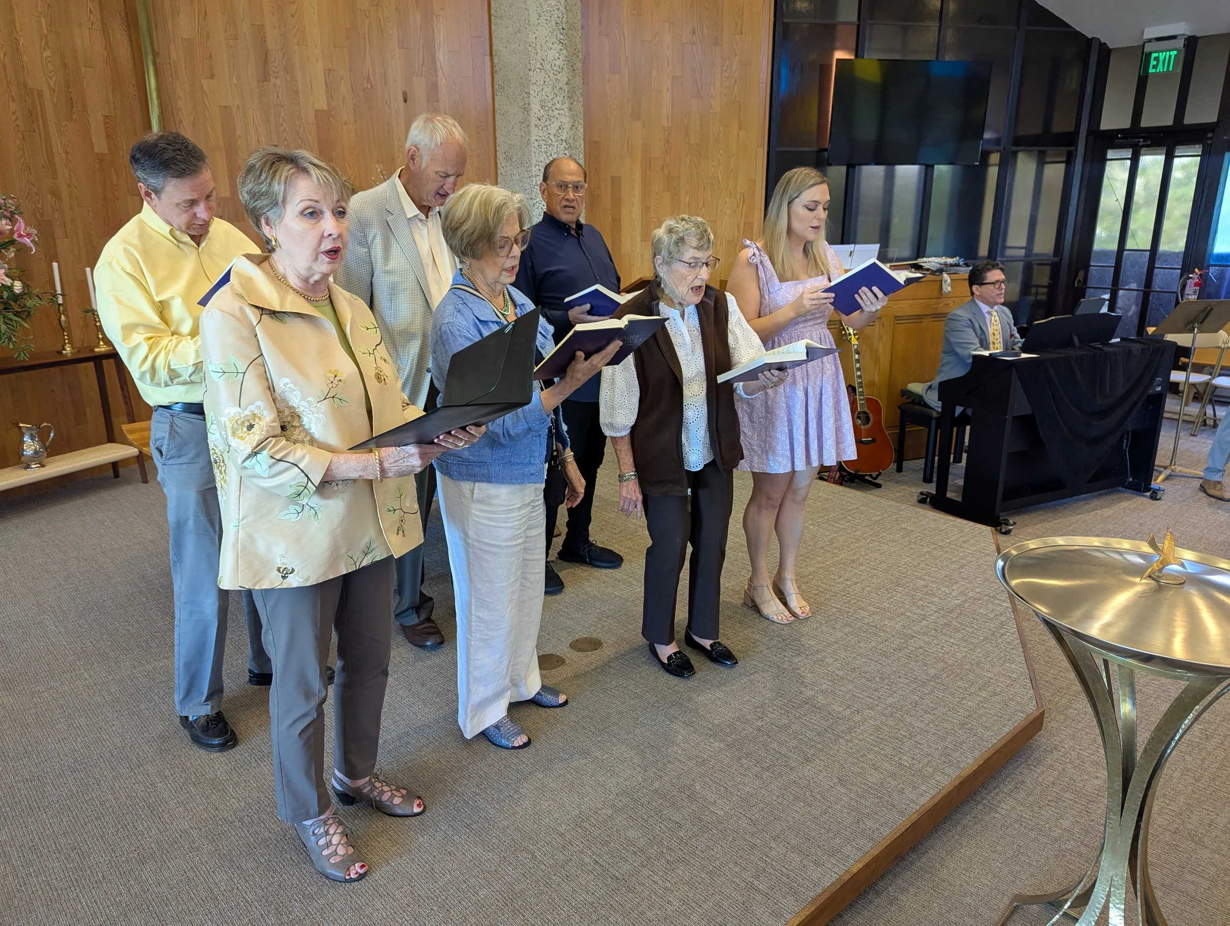 A group of people standing on a small stage, singing from songbooks, in a room with wooden walls, a keyboard, and an acoustic guitar.