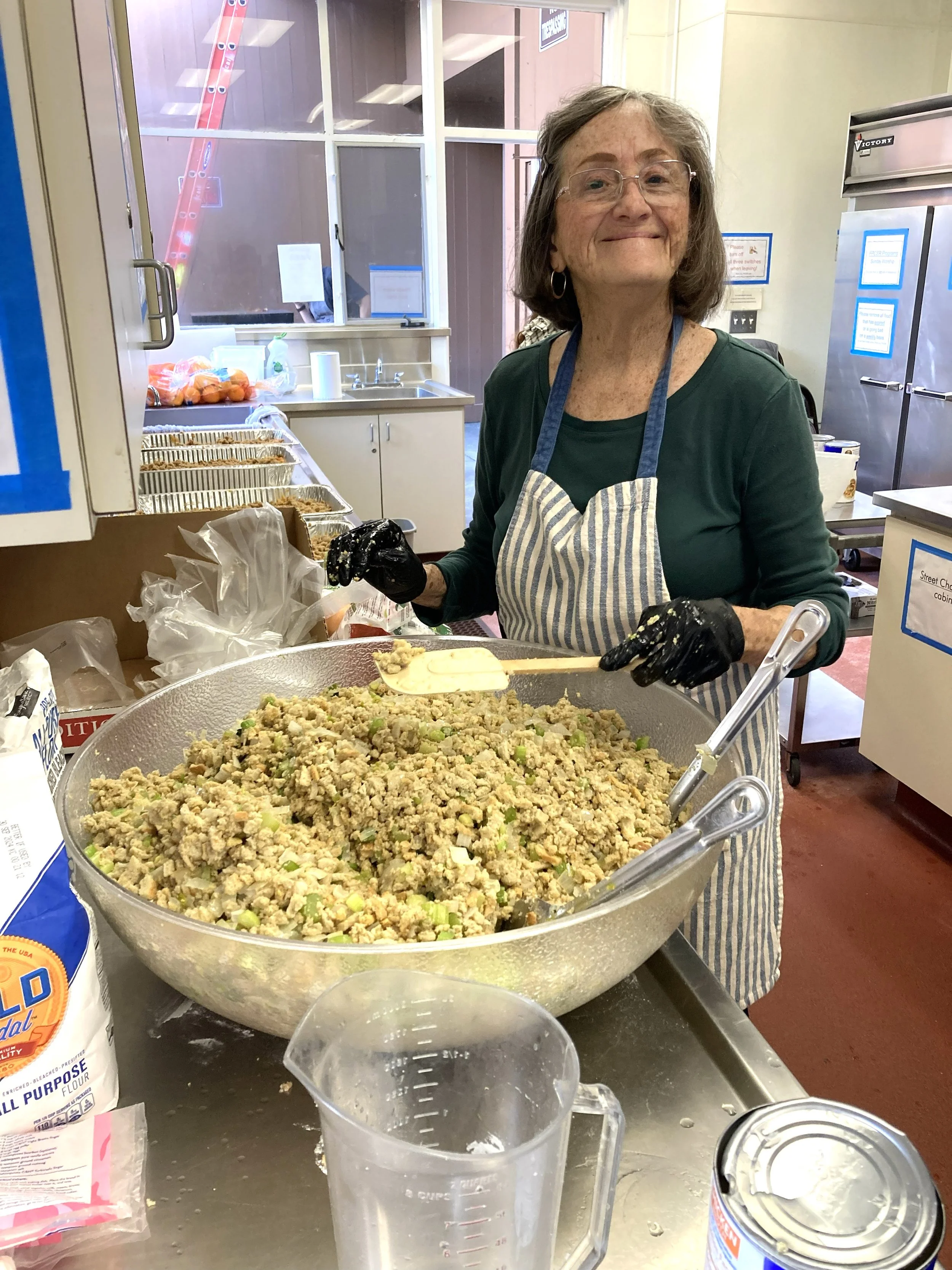 An elderly woman with glasses and earrings wearing a striped apron and gloves standing behind a large bowl of prepared food, smiling at the camera, in a kitchen or community kitchen setting.
