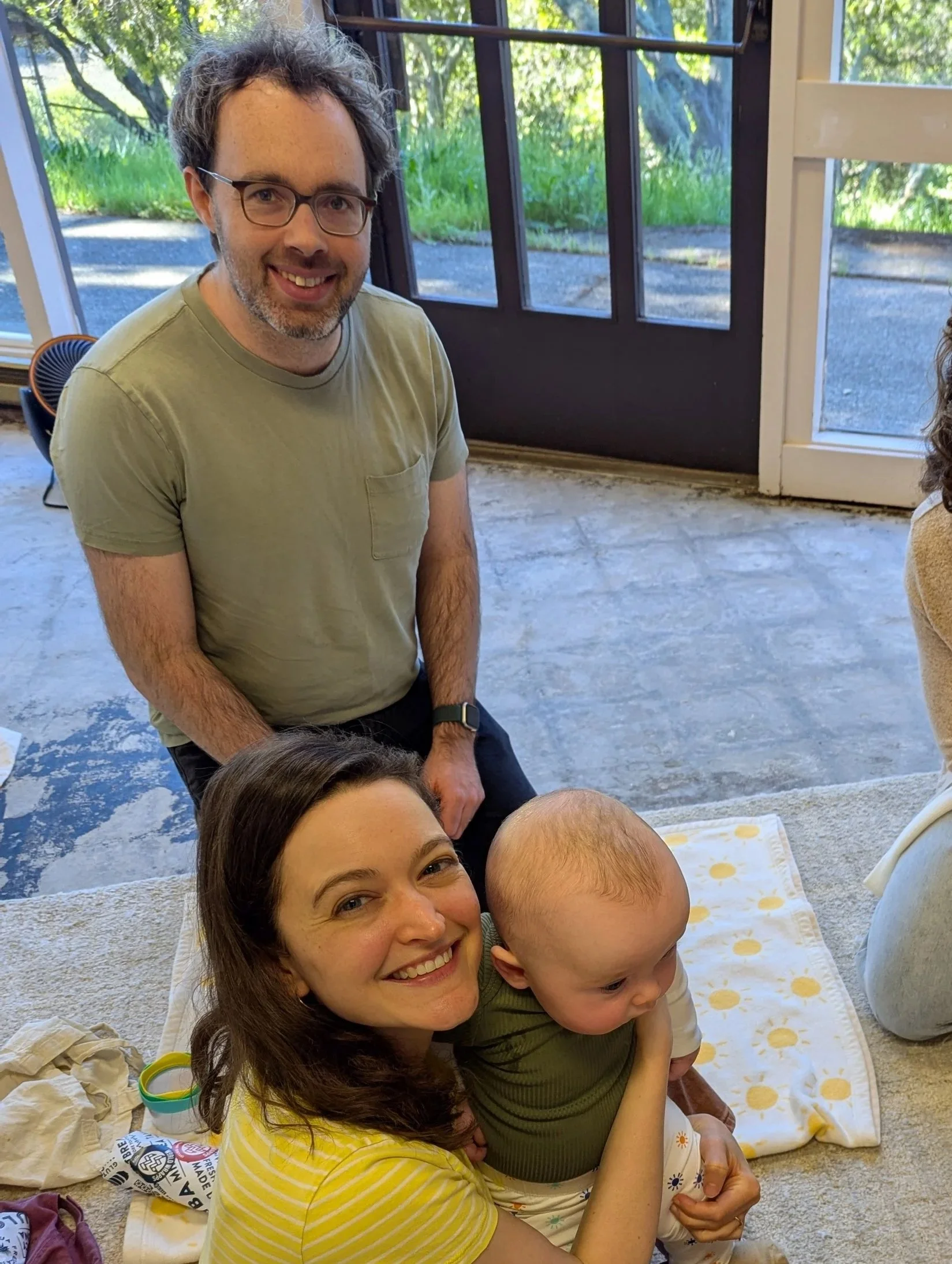 Smiling woman with brown hair holding a baby, and a man with glasses with dark hair and beard standing indoors near a door with glass panes and a green outdoor background.