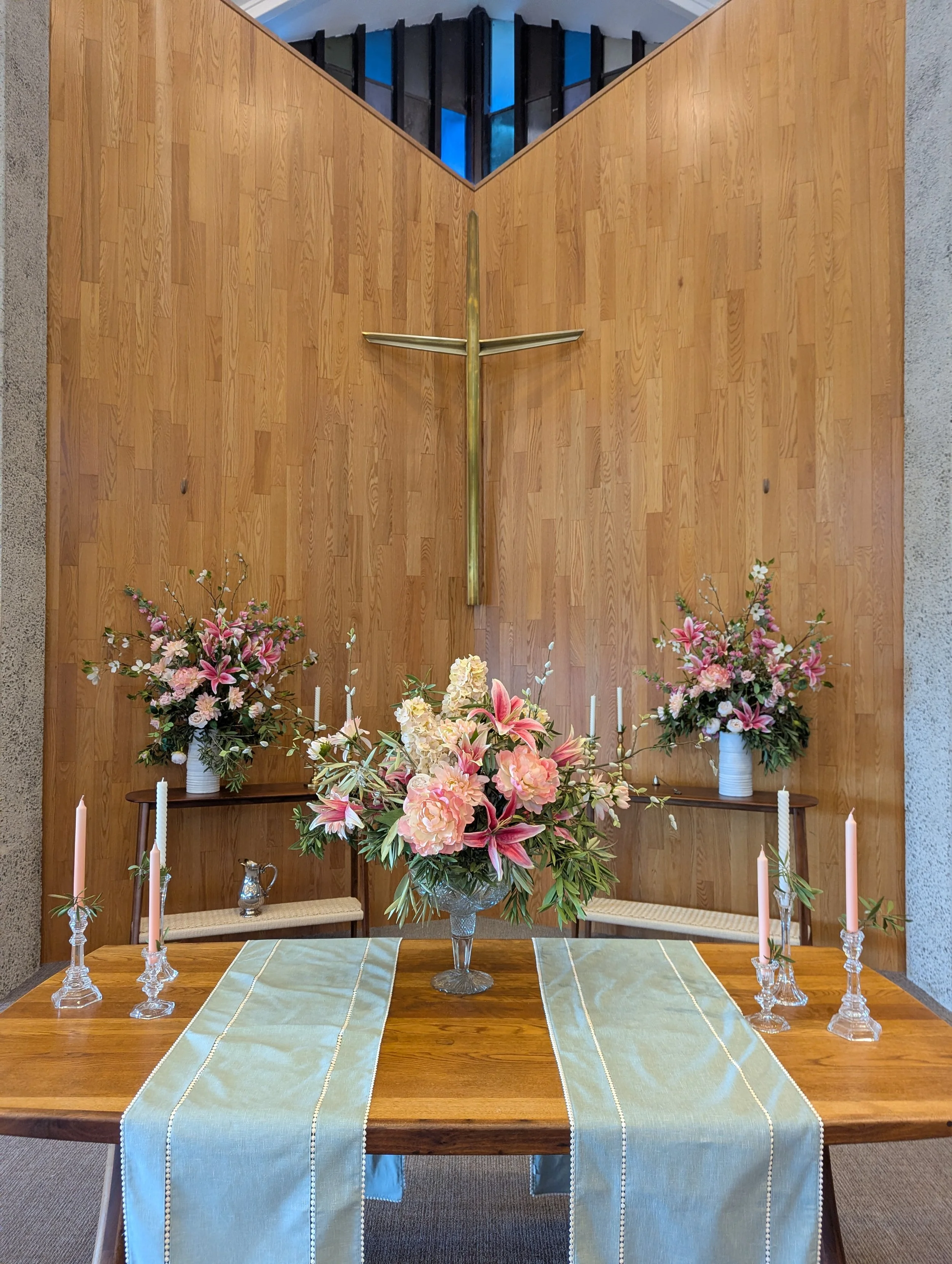 A communion table decorated with three light blue cloth altar runners, a large flower arrangement with pink lilies and white hydrangeas, surrounded by four pink candles in glass holders, and two small chairs on each side.