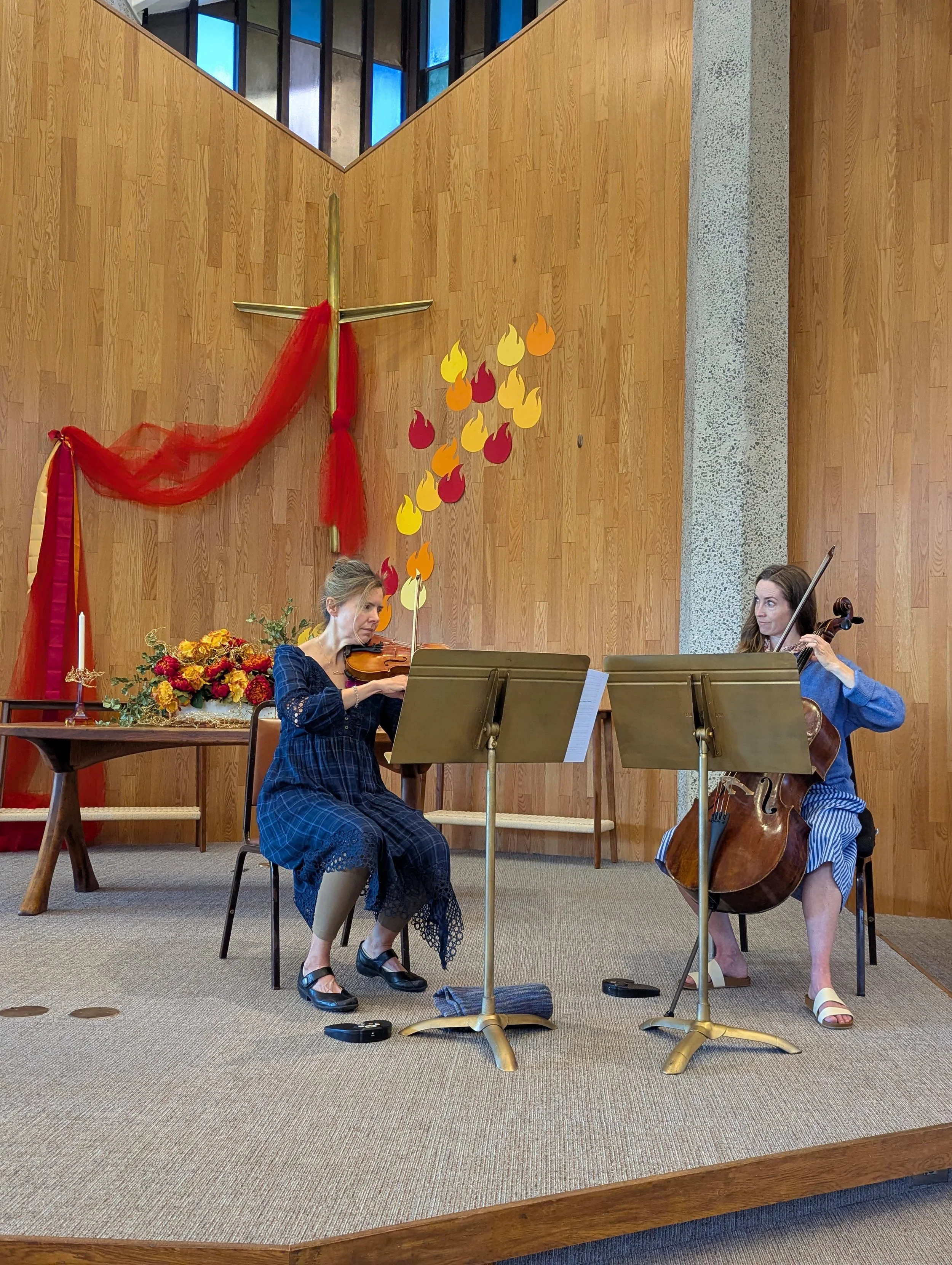 Two women playing a violin and cello during a worship service on a small stage with decorated wooden walls and a floral arrangement.