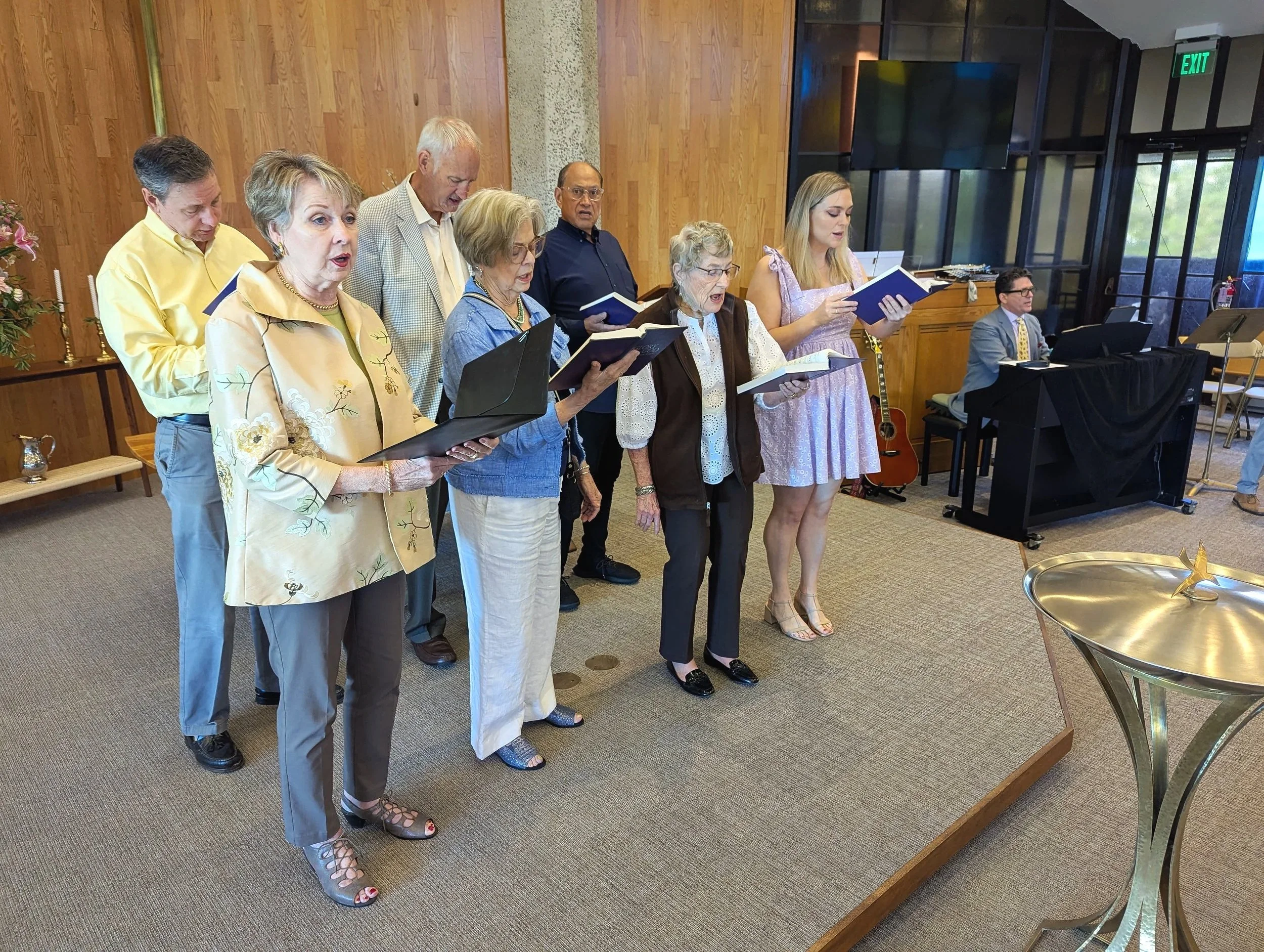 Ten people, mostly elderly, are standing in a row inside a wooden-walled room, singing from hymn books and a laptop, with a man playing a piano to the right.