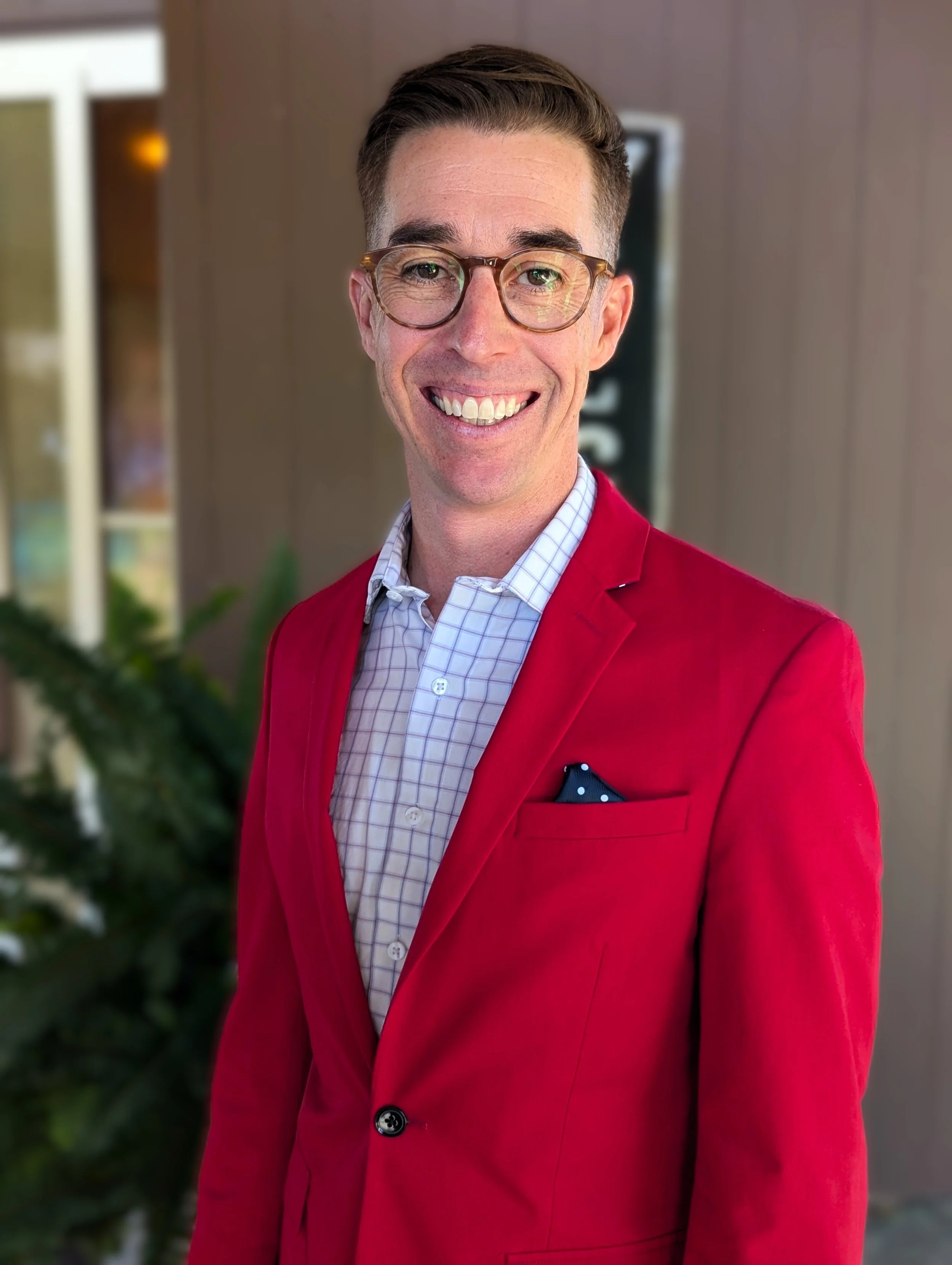 A smiling man with glasses wearing a red blazer and a checked shirt, standing outside with blurred greenery and building background.