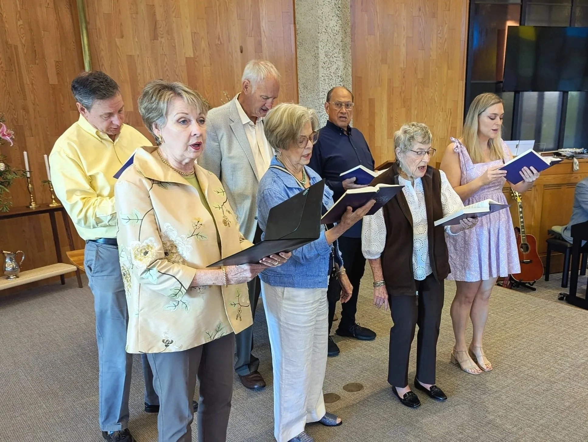Group of eight older adults standing indoors, singing from hymnals, with wooden wall and musical instruments in background.