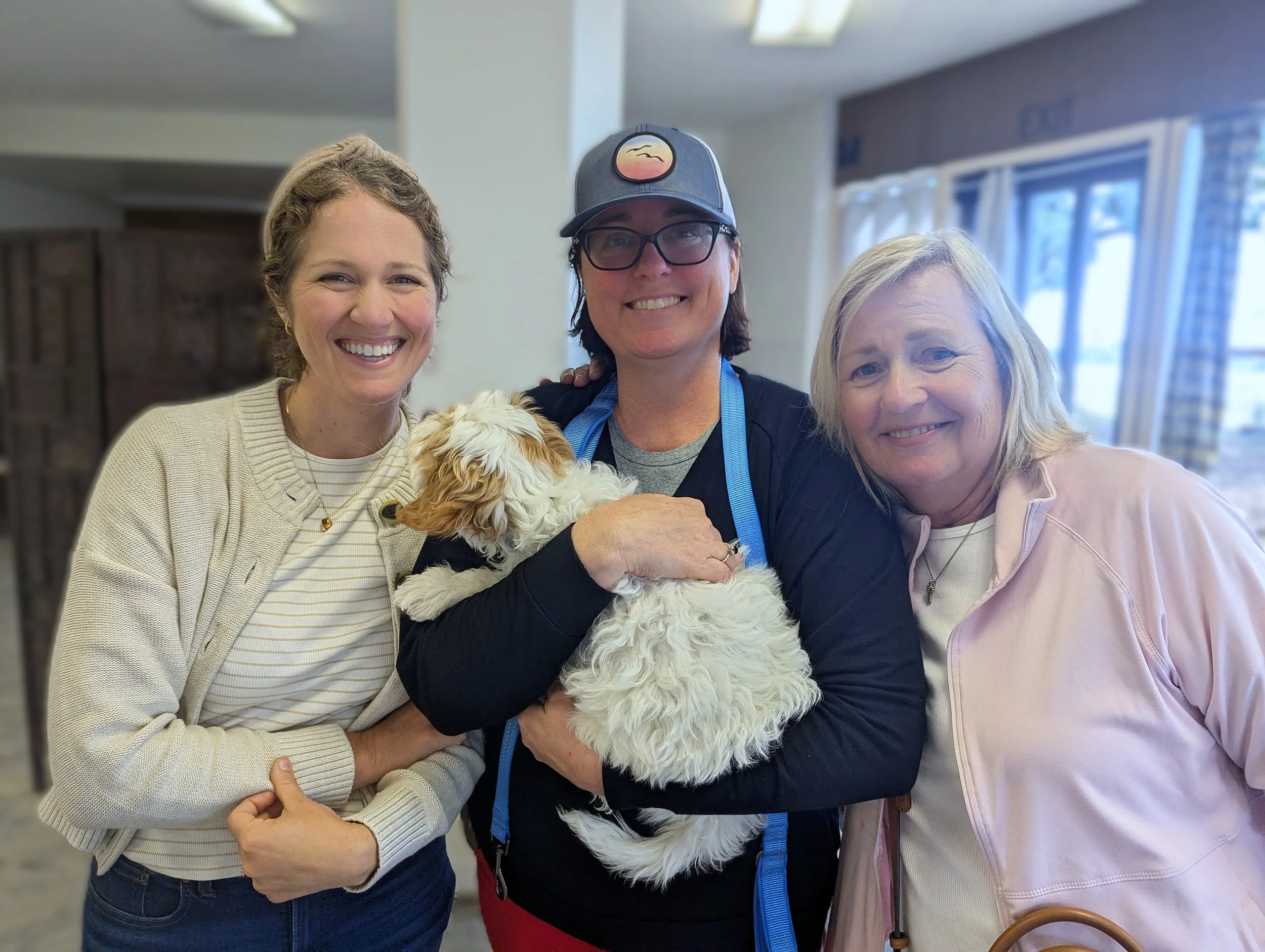 Three smiling women and a woman holding a small white and brown dog inside a room with windows and curtains.