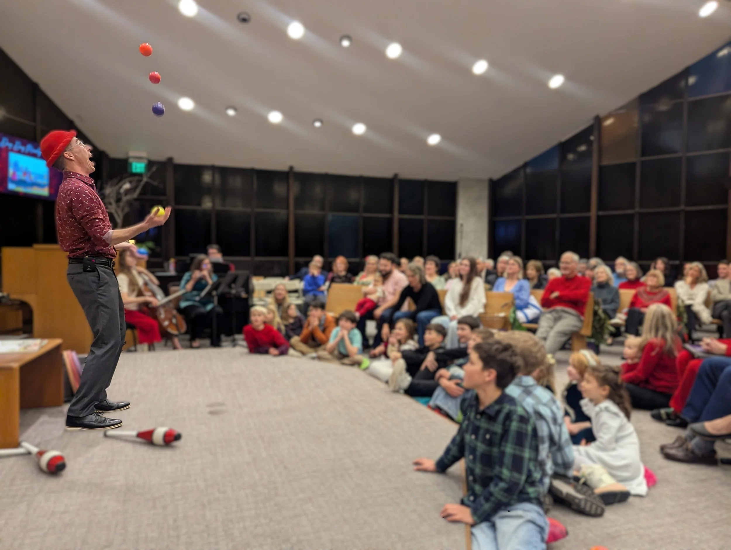 A performer juggling multicolored balls in front of an audience of children and adults seated on the floor and chairs in a modern indoor space.