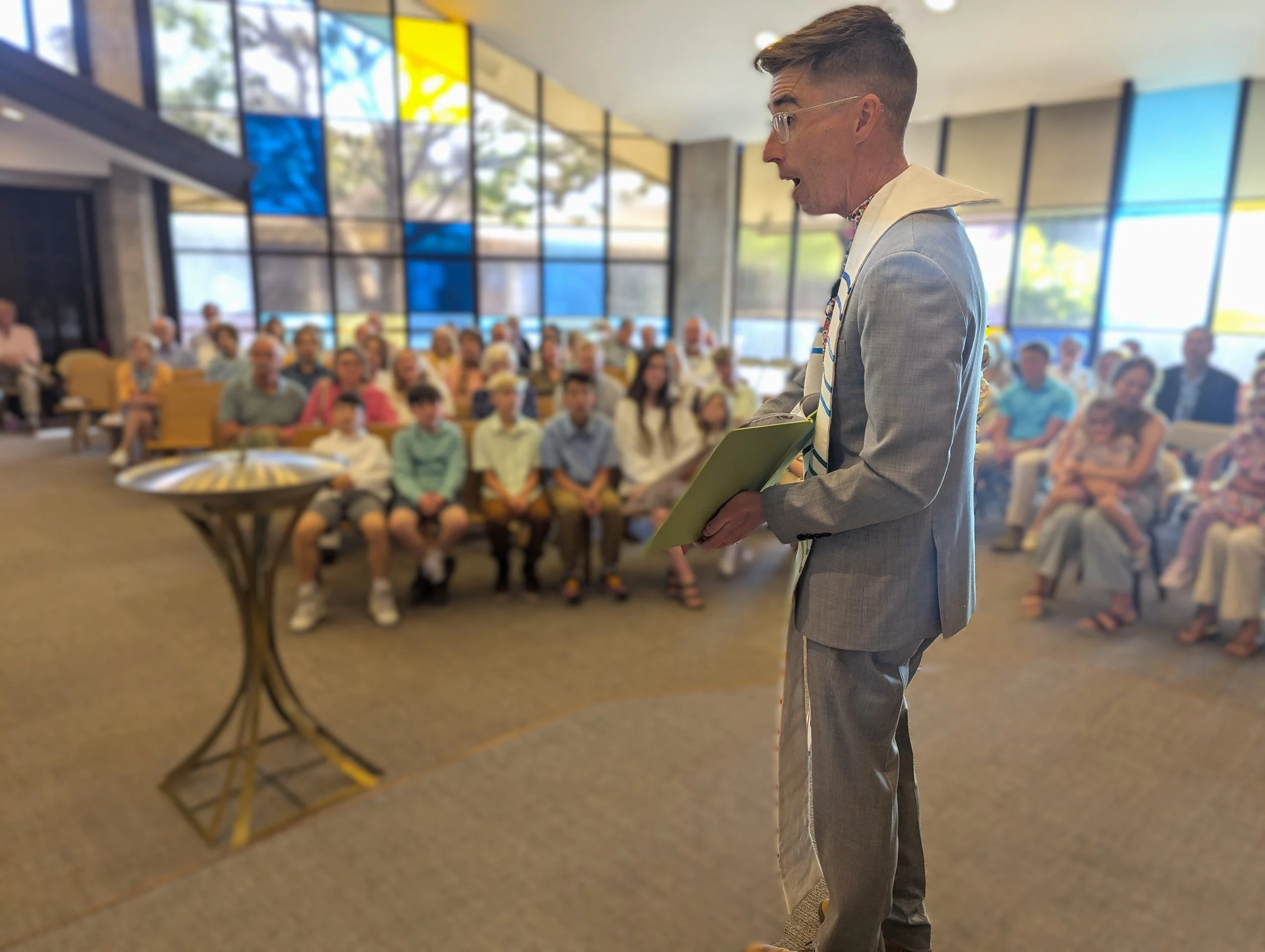 A man in a gray suit and glasses holding a children's book, reading in front of a seated audience in a modern indoor sanctuary with large colorful stained glass windows.