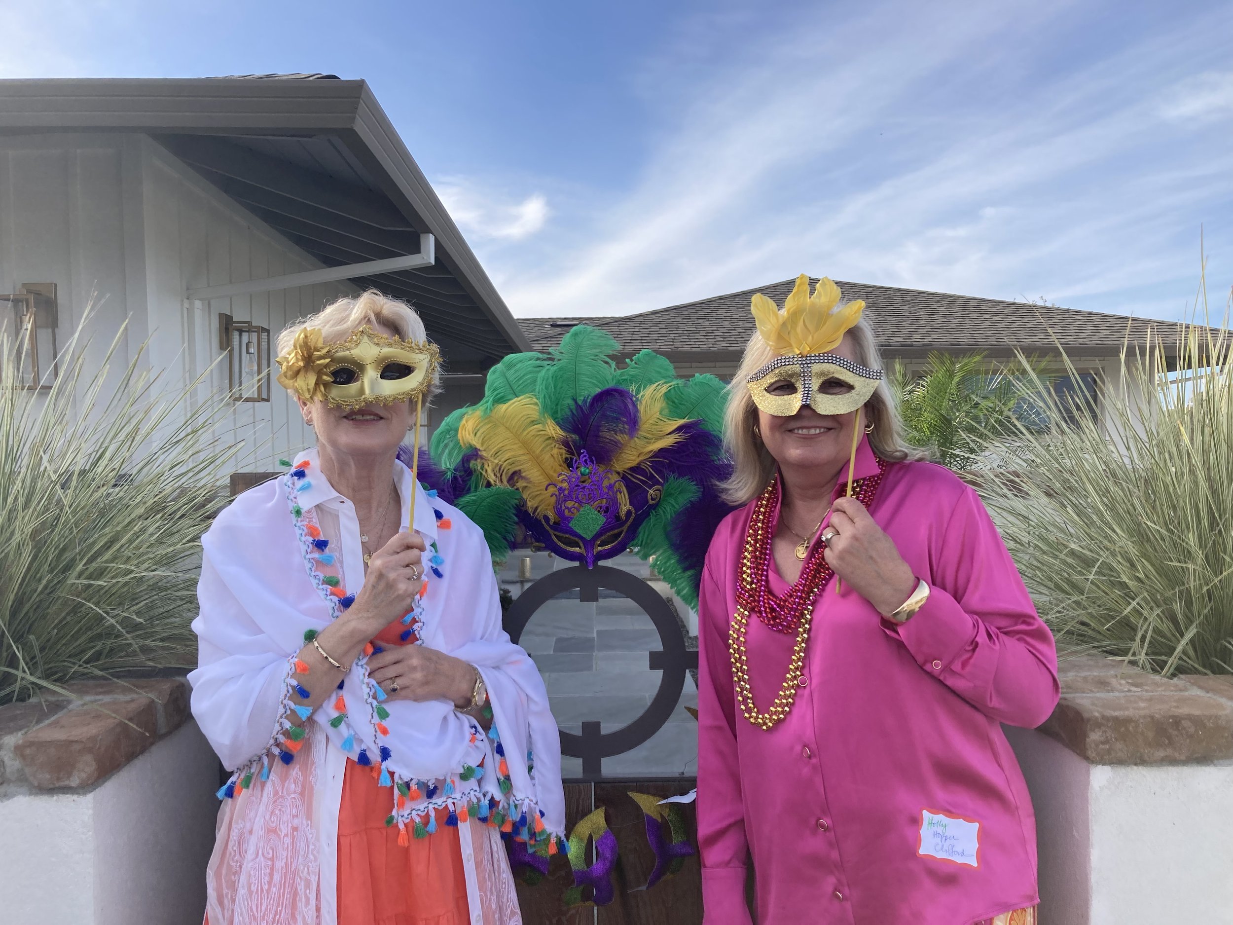 Two women wearing gold and glitter masks and colorful beads, standing outdoors in front of a house with a gray roof and blue sky, celebrating Mardi Gras.