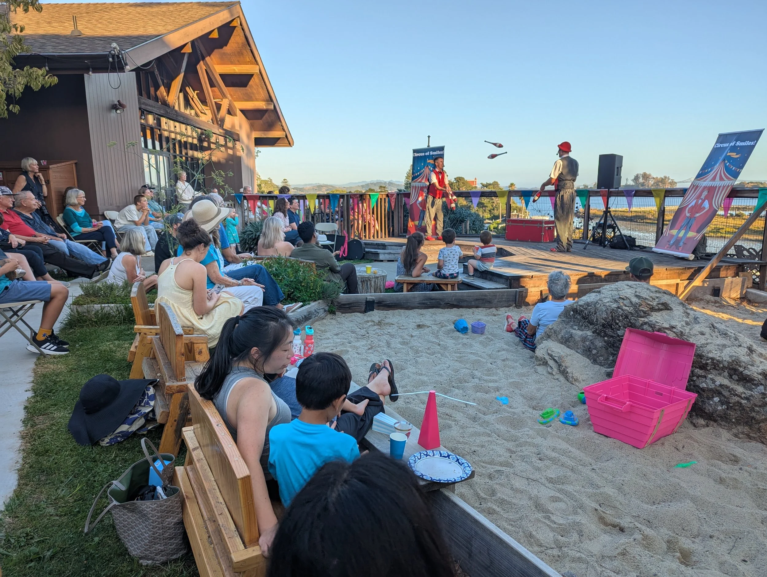 An outdoor circus show with performers on stage juggling balls, in front of an audience consisting of children and adults, with colorful banners, and a sand area with toys.