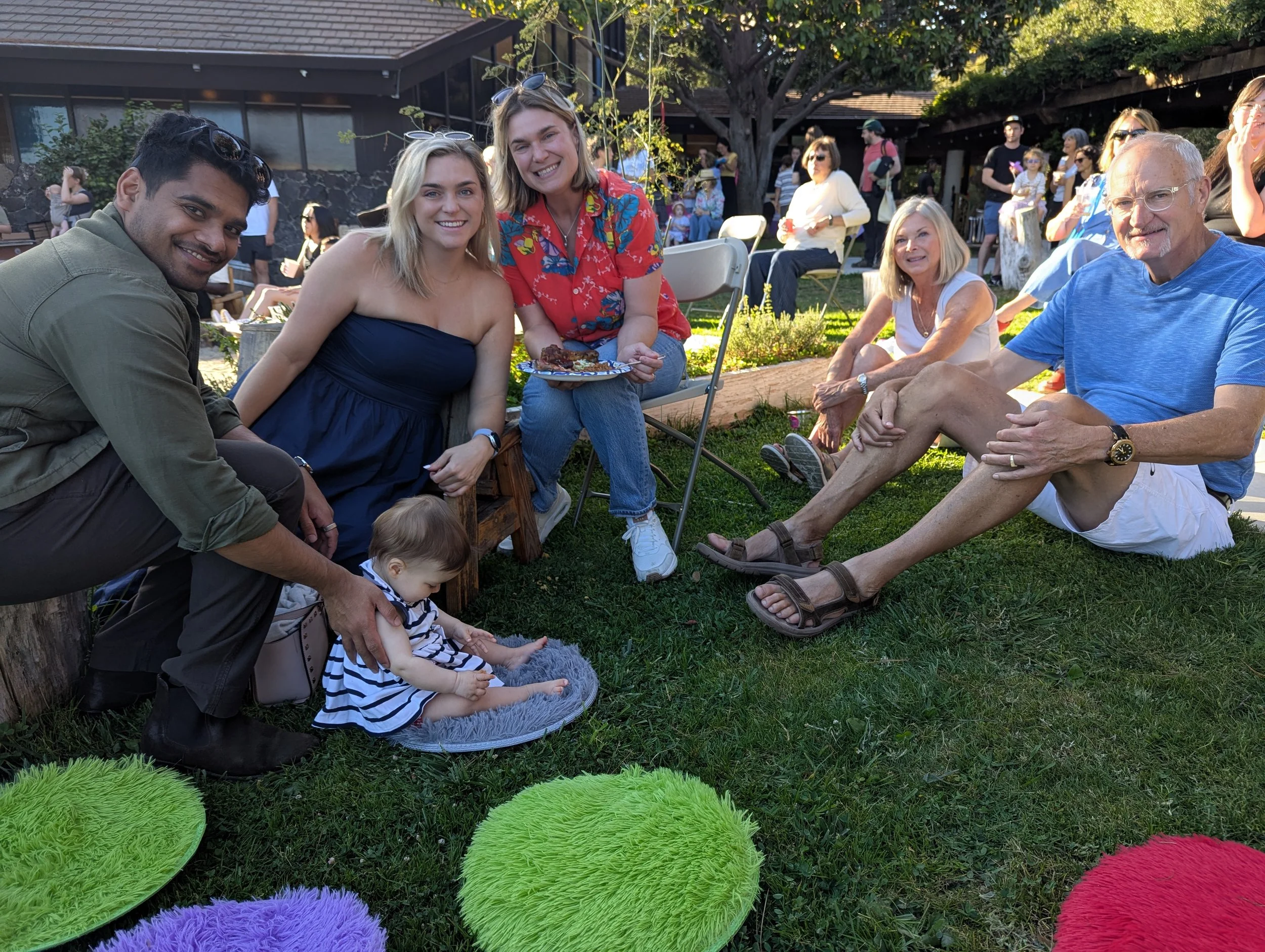 A group of people sitting outdoors on grass during a social gathering, with colorful fuzzy mats in the foreground, smiling and enjoying the event.