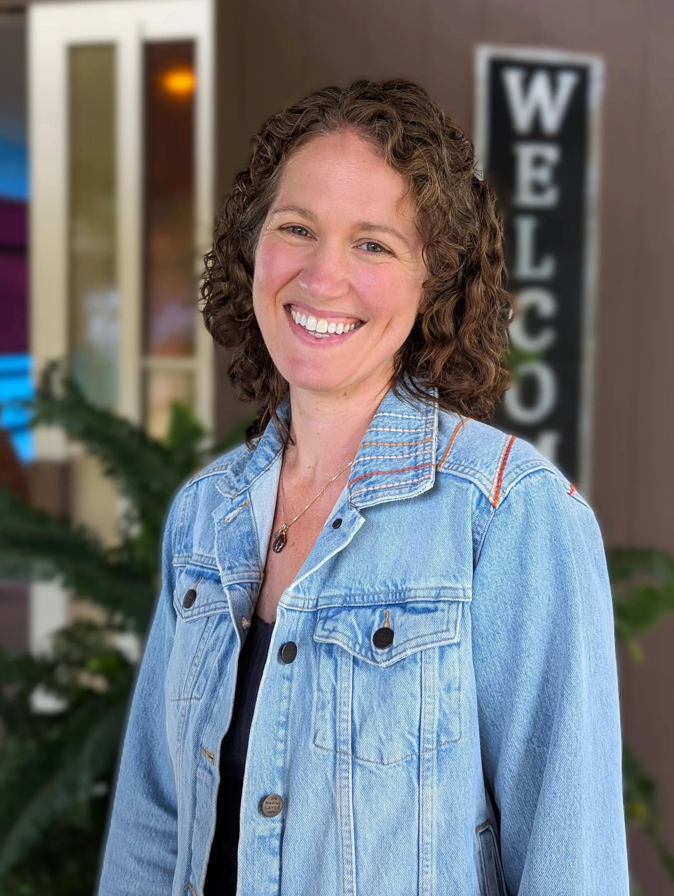 A smiling woman with curly brown hair wearing a denim jacket, standing outdoors with a blurred background that includes signs and possibly a school or community center.