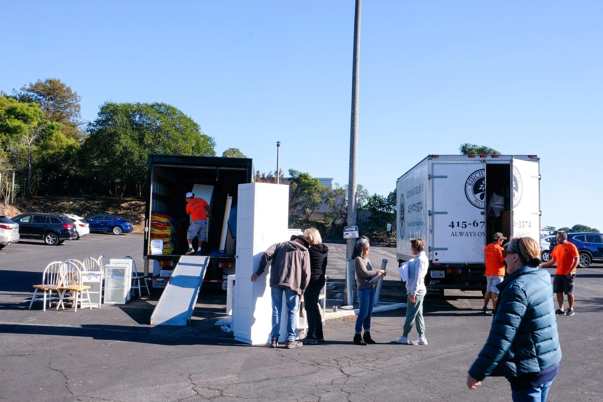 People gathered in a parking lot outdoors, possibly at an event, with two trucks and an open moving truck. Some individuals are talking, and chairs are visible on the ground. There are trees and a clear blue sky in the background.