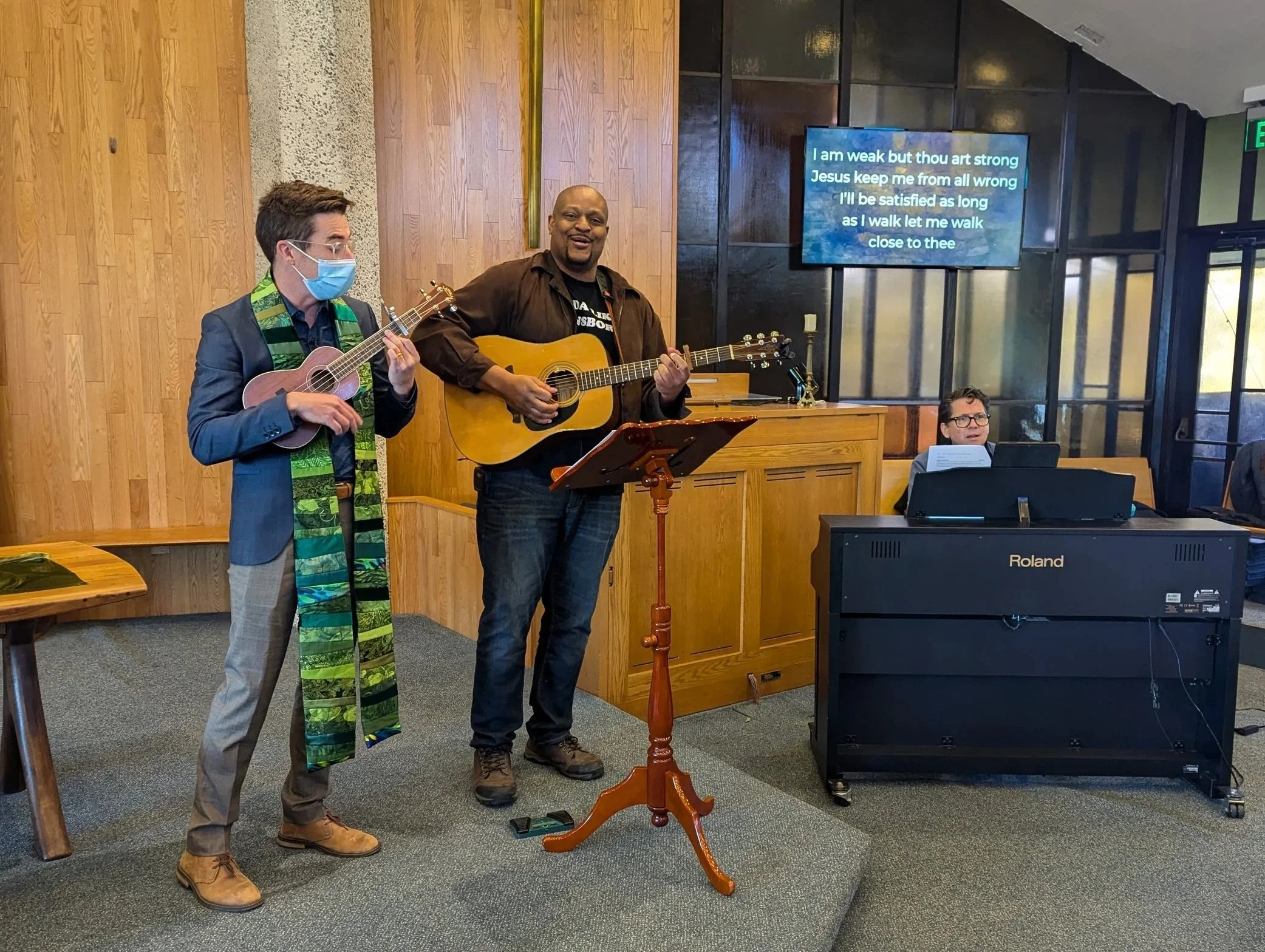 Two men playing guitars in a church or community center, one wearing a face mask, and a man sitting at a keyboard, with a large TV screen displaying song lyrics in the background.
