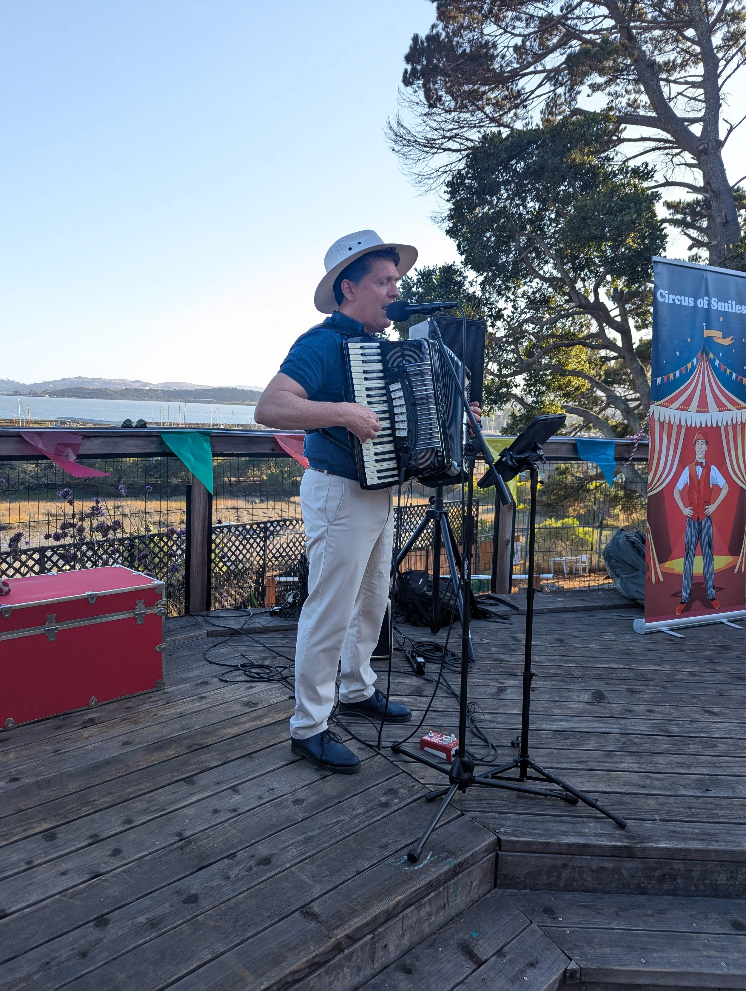 A man wearing a white hat and blue shirt playing an accordion on an outdoor wooden deck with a microphone and music stand. There is a colorful circus-themed banner on the side and a scenic view of water, trees, and hills in the background.