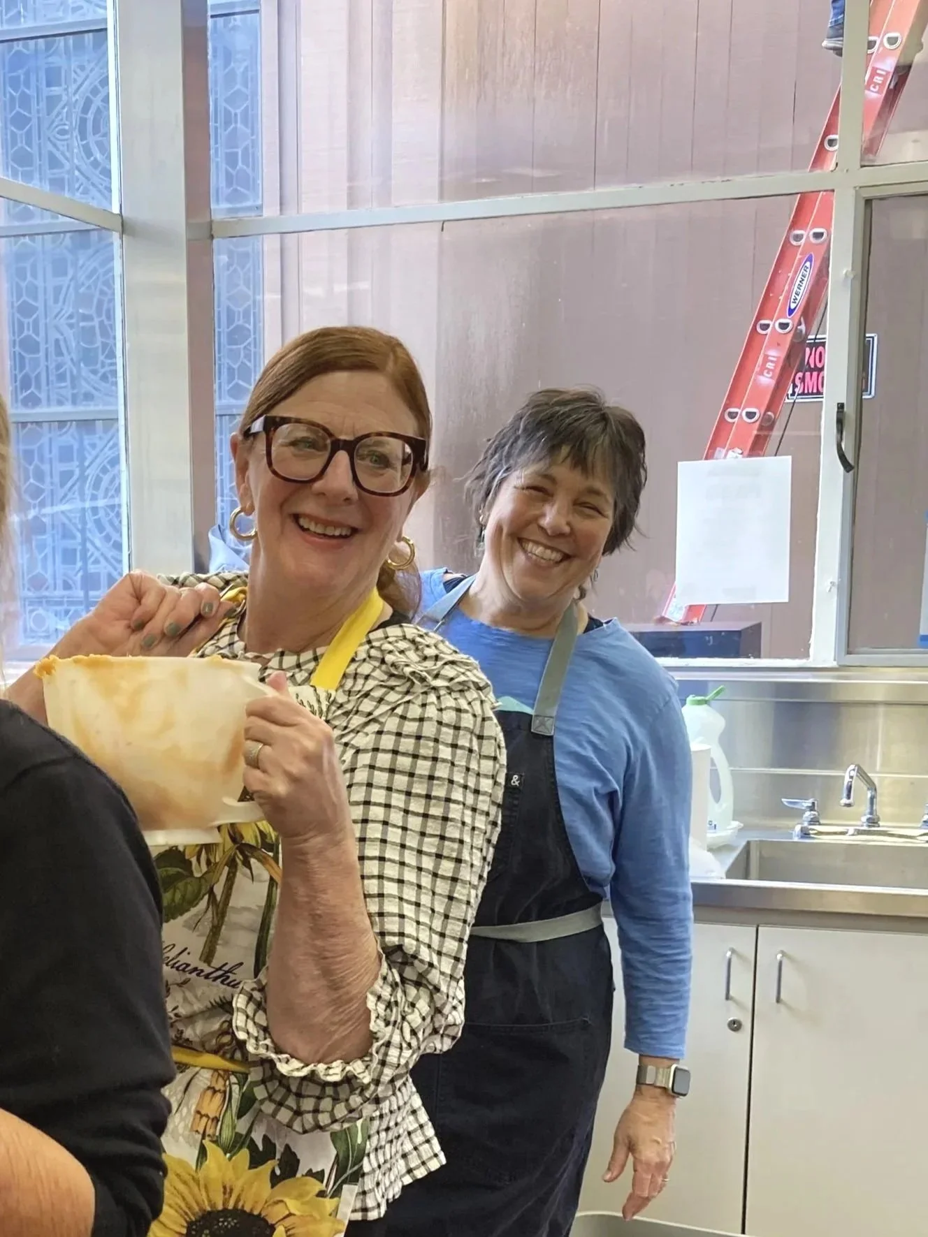 Two women smiling in a kitchen, one holding a bowl, with a window and kitchen sink in the background.