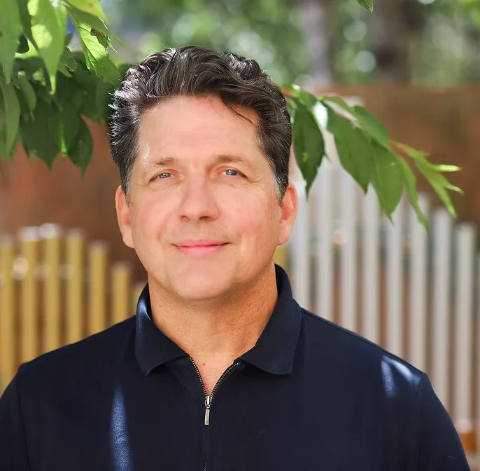 A man with dark, wavy hair and blue eyes standing outdoors with green foliage and a wooden fence in the background, smiling gently at the camera.