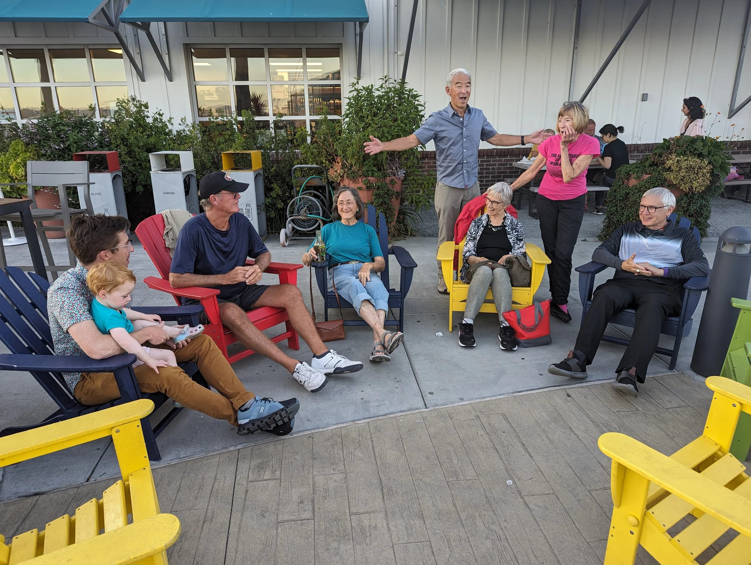 A group of people gathered outside on a patio, sitting on colorful chairs and enjoying a social moment. Two people are standing and engaging with the seated individuals, one with arms outstretched expressing enthusiasm. The setting includes potted plants, a building with large windows, and a few other people in the background.