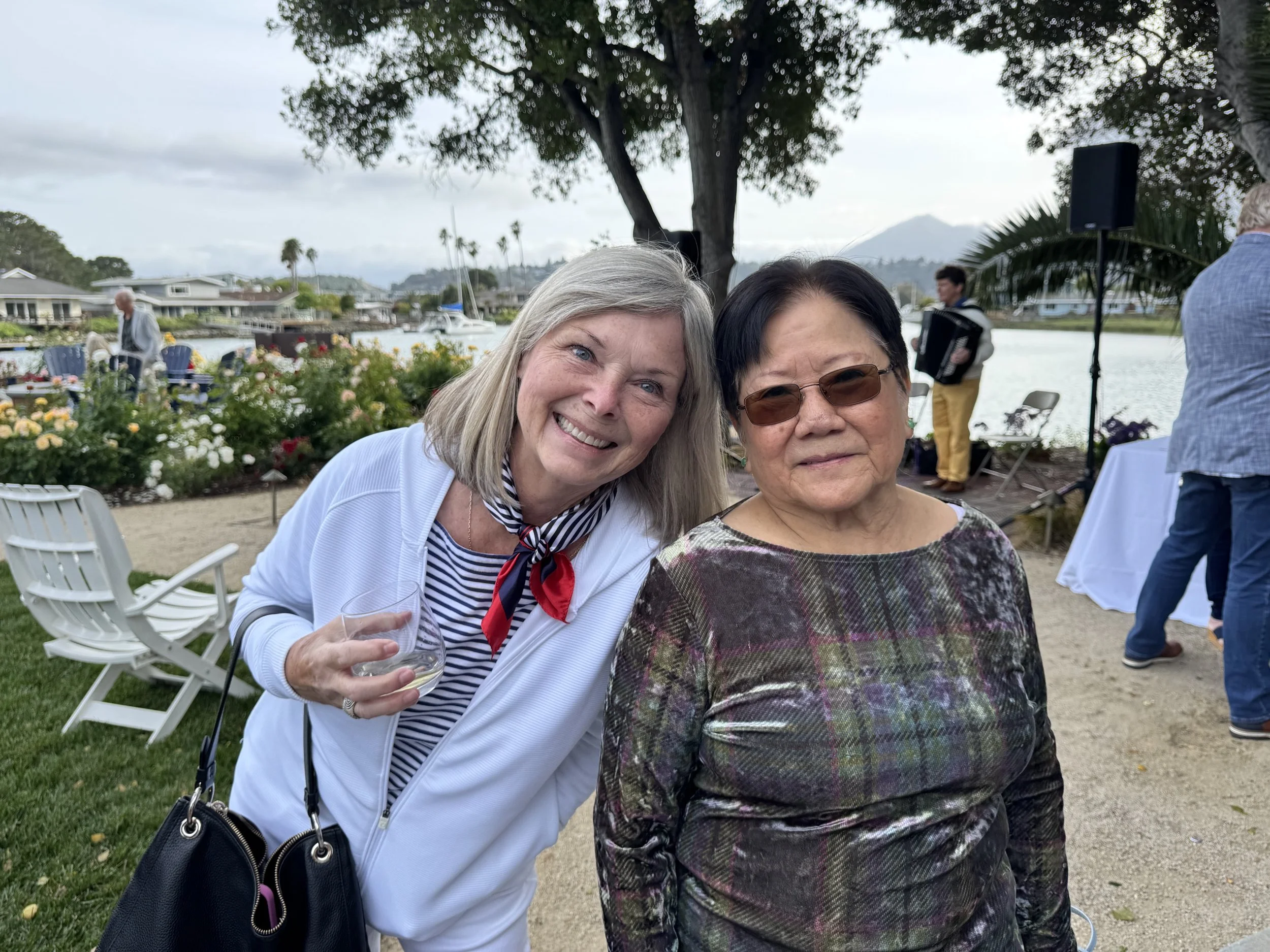 Two women standing close together at a waterfront outdoor event, smiling at the camera. One woman has gray hair and is holding a glass of wine; the other has black hair and is wearing sunglasses. In the background, there are people, trees, boats, and