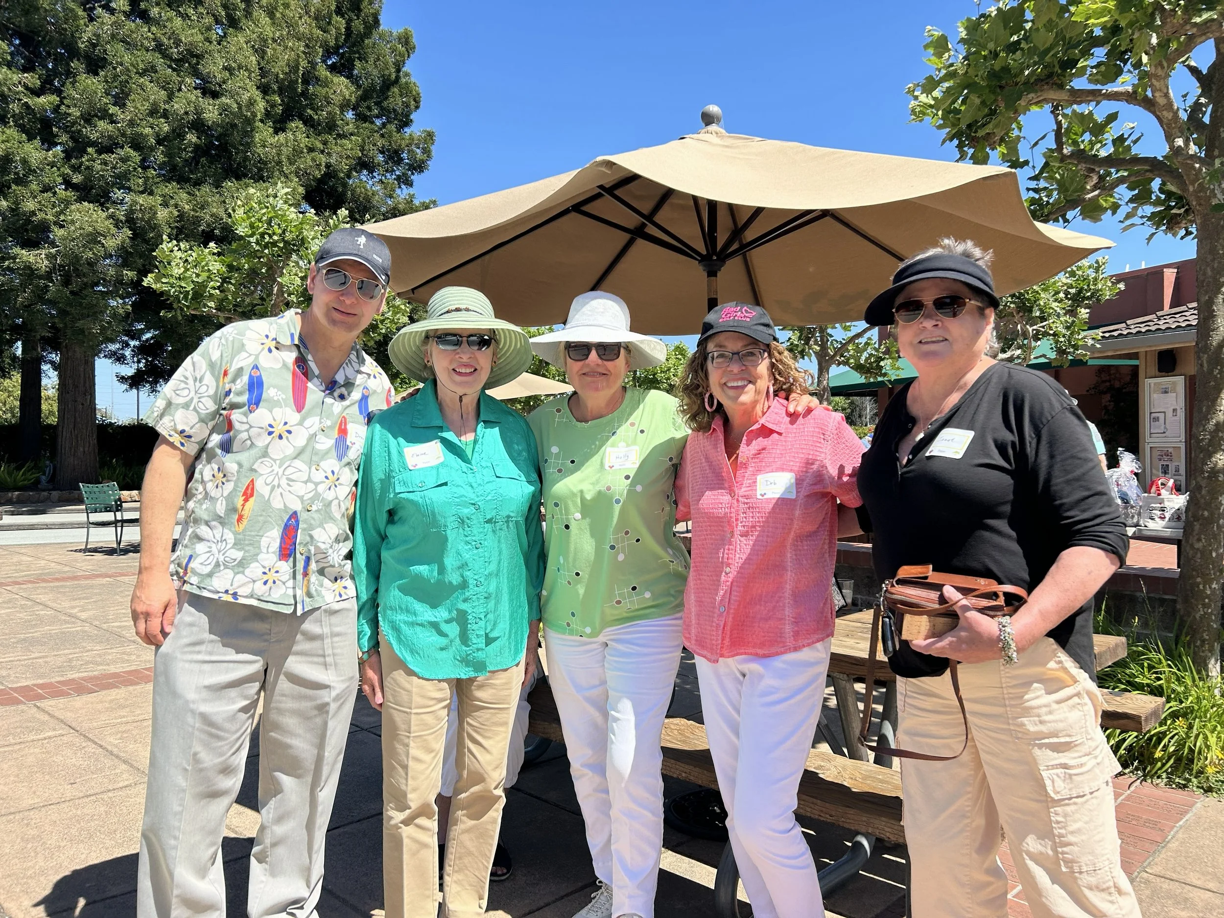 A group of adults standing outdoors under a large beige patio umbrella, posing for the photo. They are dressed in casual summer clothing and sunglasses, with some wearing hats. The background shows trees, a building, and a blue sky.